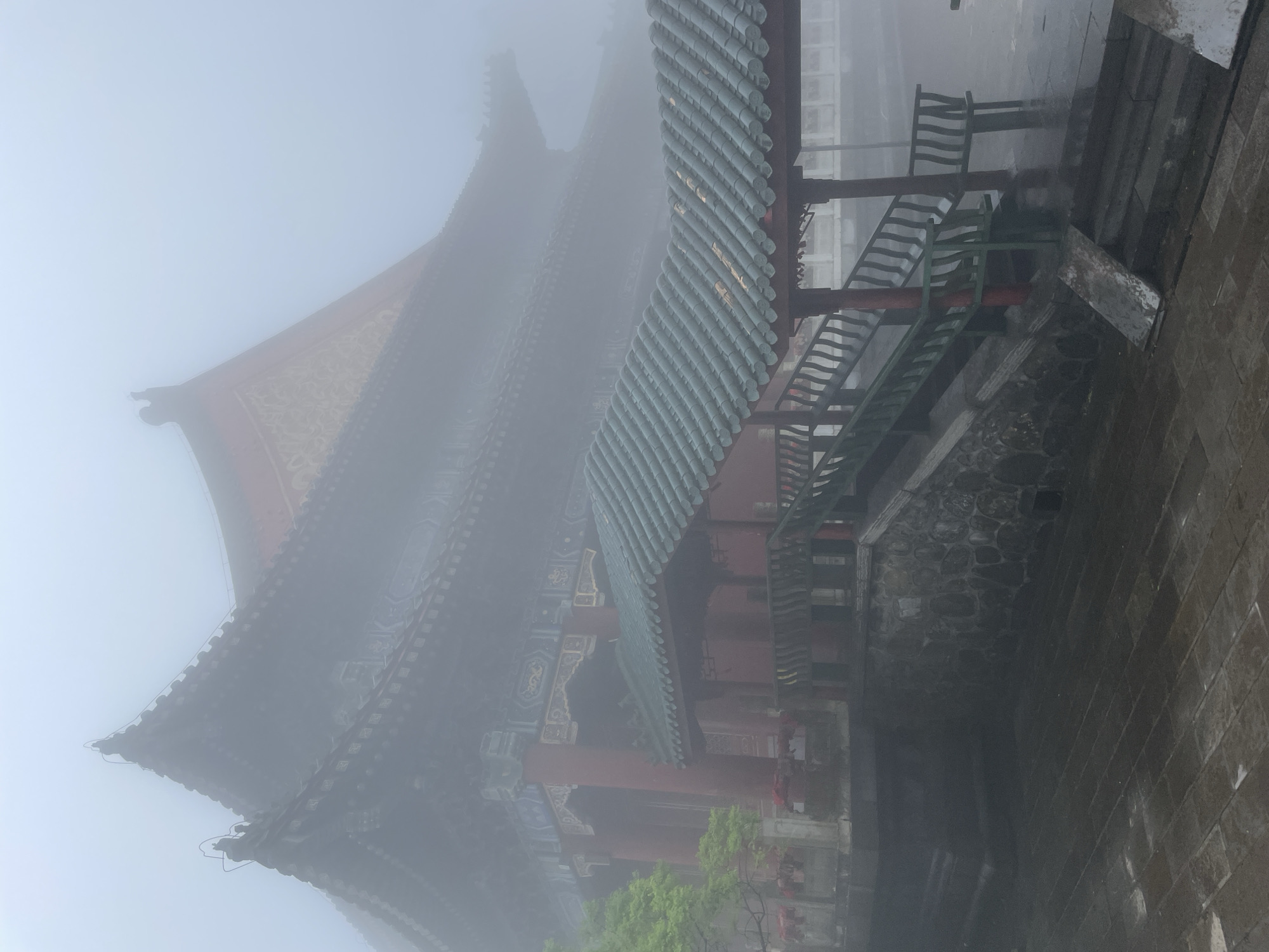 The image shows a traditional Chinese building shrouded in heavy fog. The structure features layered, tiled roofs, red pillars, and intricate decorative details. Adjacent to the building is a set of stairs and a stone wall. The scene is located in Yongding District, China.