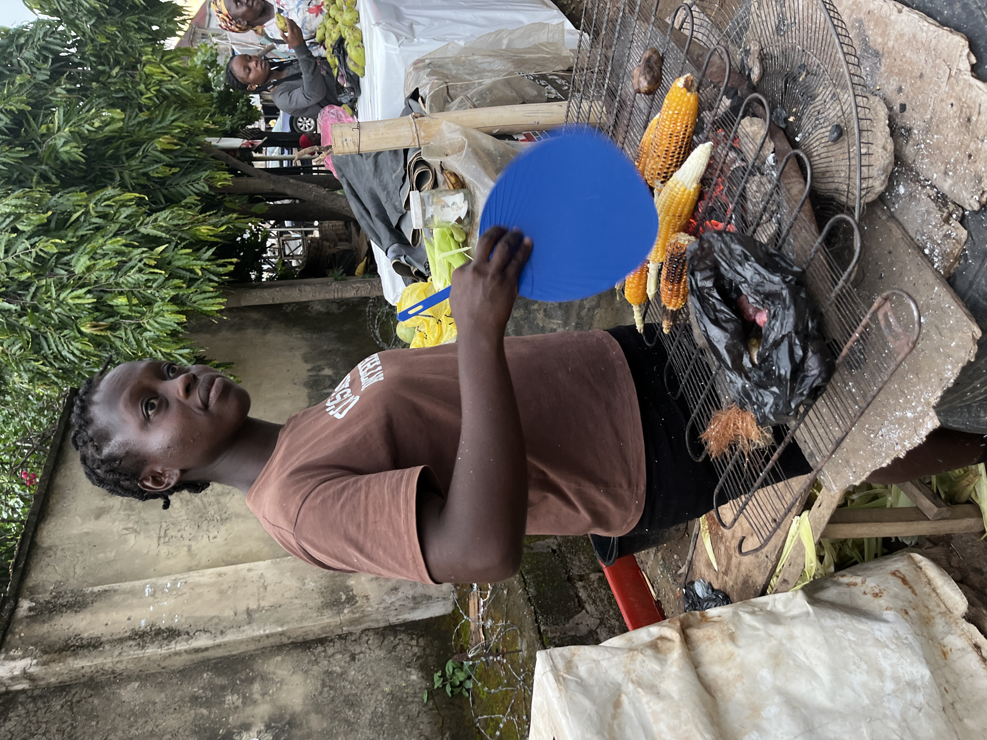 A young woman is fanning corn cooking on a grill with a blue fan. The woman is wearing a brown t-shirt and black pants. The corn is on a metal grill over hot coals. There are several ears of corn on the grill, some of which are partially cooked. In the background, there are other people and a table with fruit. The scene appears to be outdoors, possibly at a market. The environment suggests an urban setting, consistent with a market in Abuja, Nigeria.