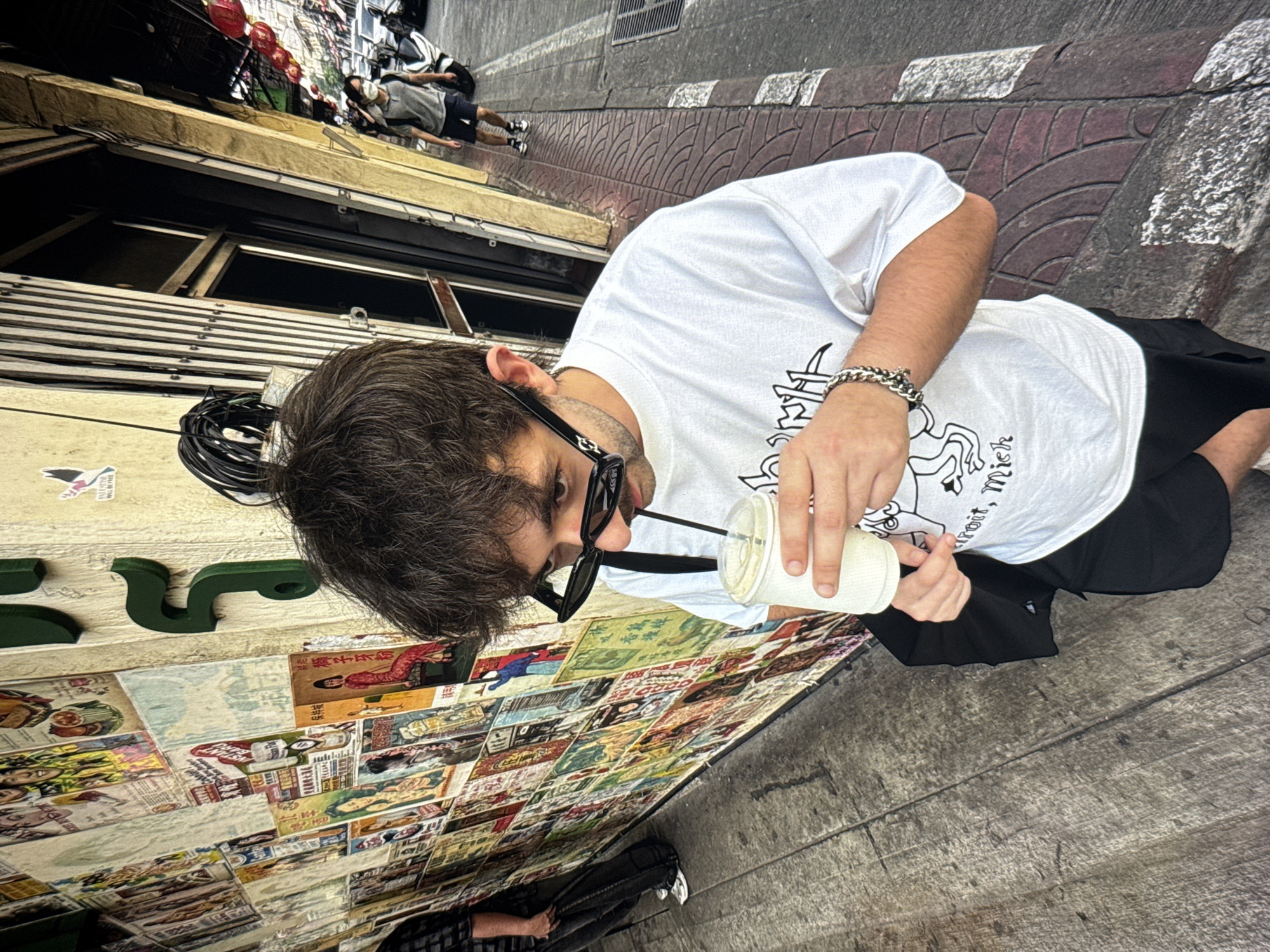 The image shows a man in a white t-shirt and black shorts standing on a sidewalk in front of a wall covered in posters. He is holding a cup with a straw and drinking from it. He is wearing black sunglasses and a silver bracelet. In the background, there is a street with buildings and a few people walking. The man's hair is brown and messy. The posters on the wall are colorful and appear to be advertisements or movie posters. The sidewalk is made of cobblestones. The lighting suggests it is daytime. The location is not Moscow, Russia.