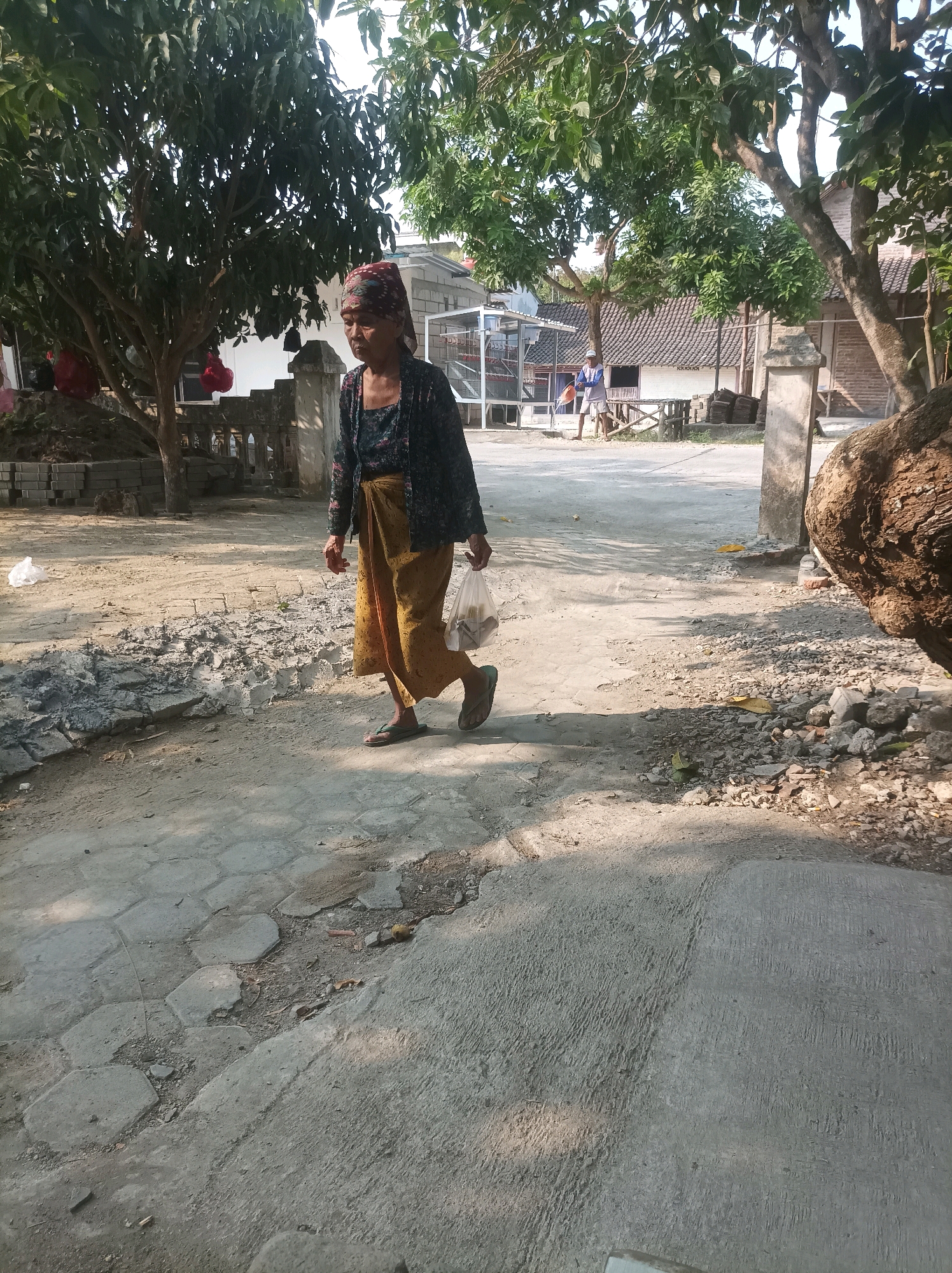 In CANGA'AN, Indonesia, an elderly woman with a patterned headscarf walks along a paved path. She is wearing a dark patterned blouse and a long, mustard-yellow skirt. She carries a transparent plastic bag in her right hand. The path is cracked and uneven, with some areas showing loose gravel and dirt. Trees with dense green foliage are visible on either side of the path, and in the background, buildings with tile roofs and a fence can be seen. The overall scene suggests an outdoor, rural or semi-urban environment in Indonesia.