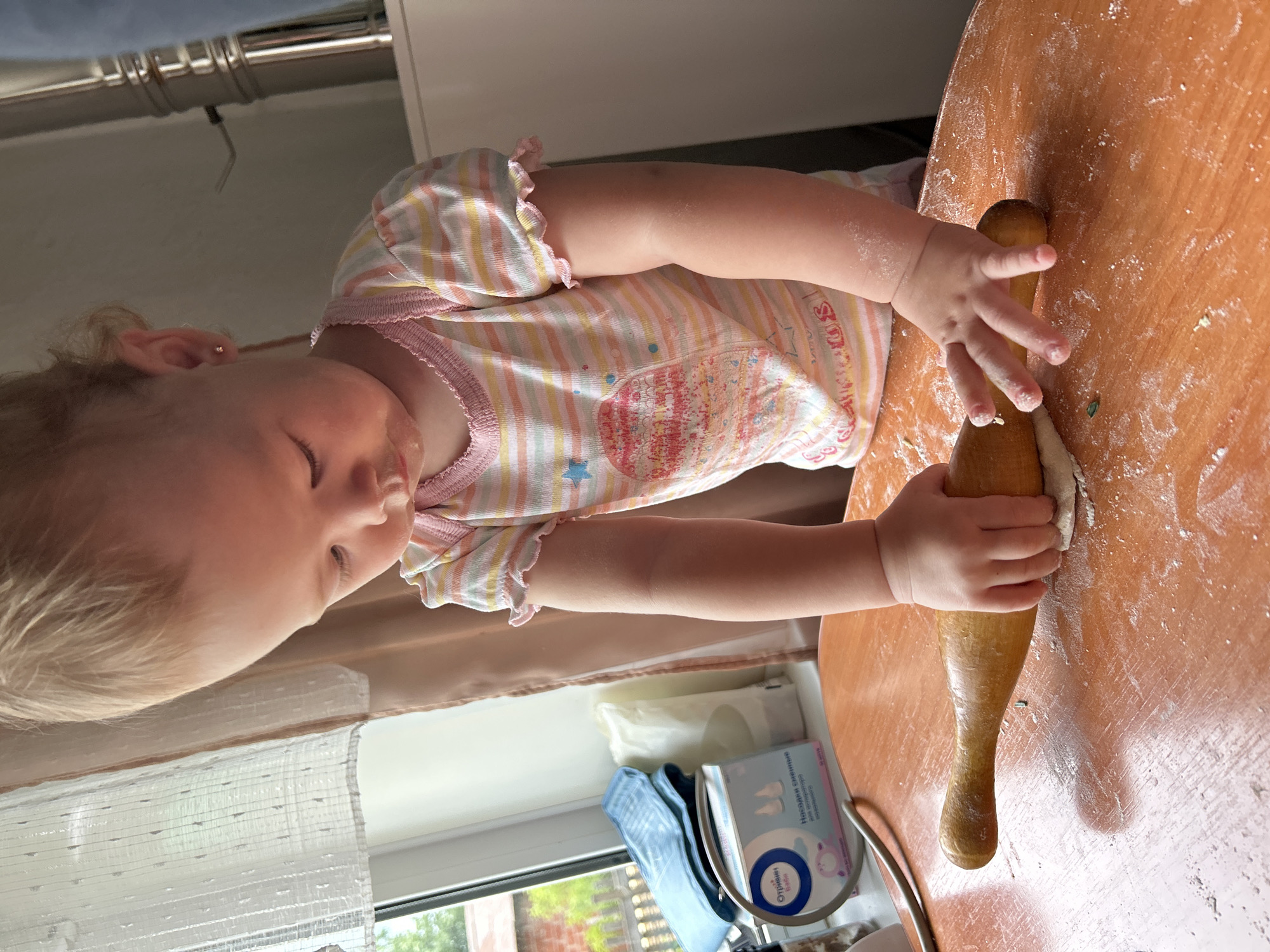 An over-the-shoulder, medium shot captures a young Caucasian toddler, with blonde hair, engaged in rolling out dough on a wooden table. The toddler, wearing a striped t-shirt with ruffled sleeves and a pink neckline, is focused on the task, their hands gripping a wooden rolling pin. Flour is scattered across the orange-toned table, indicating the ongoing activity. In the background, a window with sheer curtains reveals a glimpse of greenery and trees, suggesting an outdoor environment. To the left, on the windowsill, are several items including a box of "Отличные детки" (Excellent Kids) brand nasal aspirator replacement tips and some folded blue material. The scene appears to be set in a domestic environment, possibly a kitchen or dining area. The image is taken from a slightly elevated perspective. The location is not specified in the image itself, but if it were in Азинский, Russia, it would be a home within that city.