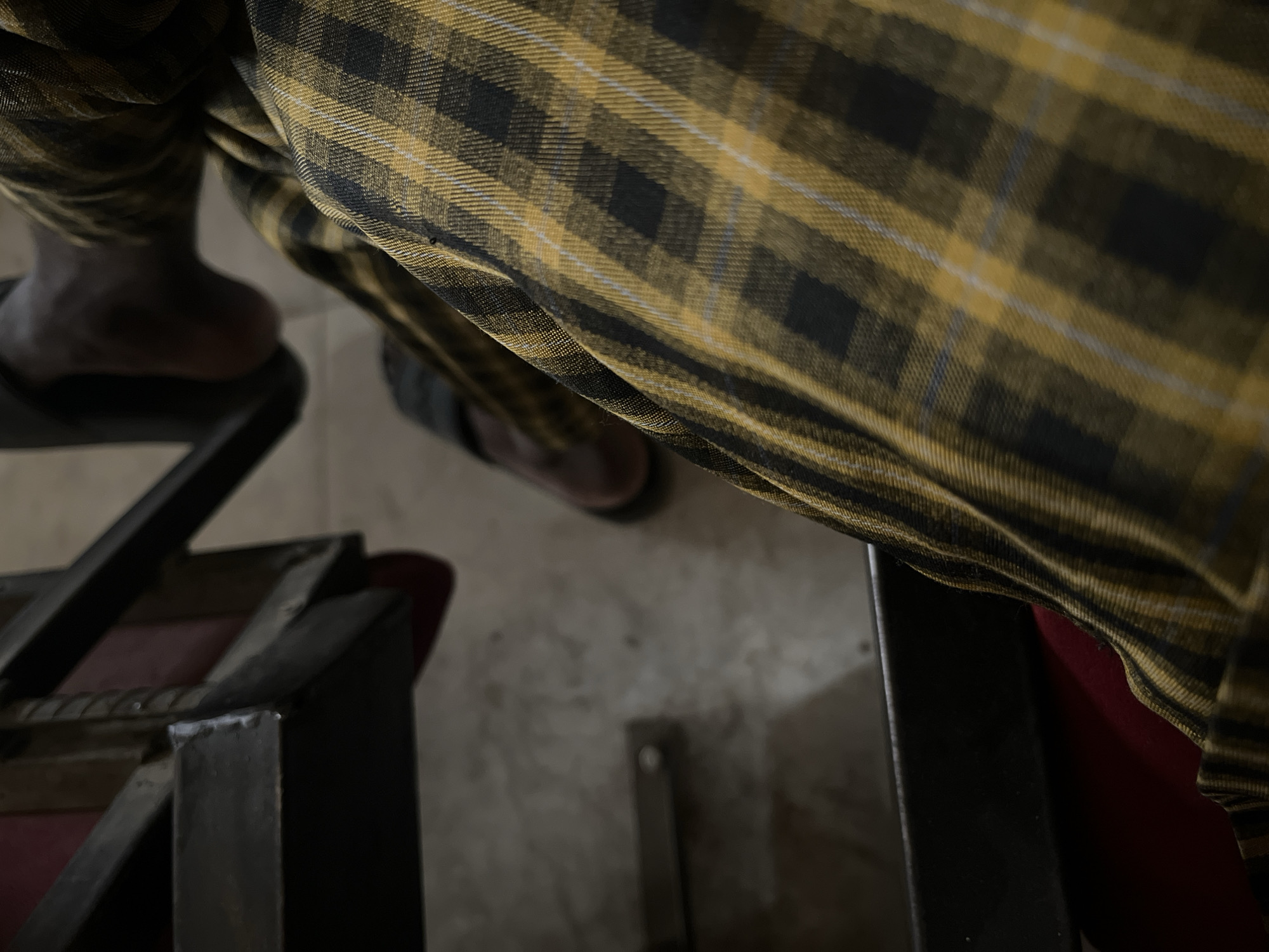 The image depicts a person wearing plaid pants in a seated position. Their dark-skinned feet are visible, one resting on the floor and the other in a black sandal atop a metal chair. A second pair of shoes is visible on the floor. The surroundings include a metal chair frame, a red seat cushion, and a grey-toned floor. The location is specified as Baganakwo, Nigeria, although there are no visual cues to confirm this.