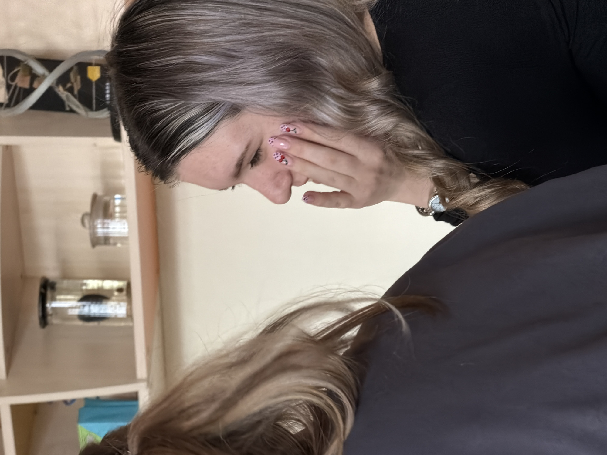 The image shows a woman with light blonde and grey hair, wearing a black top, touching her face with her hand. Her nails are painted with a cherry design. In the background is a light wooden shelf with glass jars containing dark objects. The scene takes place indoors, possibly in a salon or treatment room. There is no evidence in the image to suggest the location is in Panagyurishte, Bulgaria.