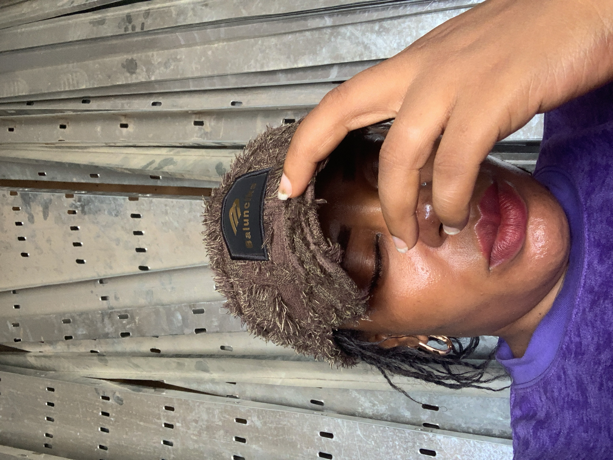 A person wearing a brown, fringed beanie with a black label is holding their hand near their face. They have dark skin, dark hair pulled into braids, and are wearing a purple top. The background consists of multiple parallel metal racks. There is no information to identify that it is Shaba, Nigeria.