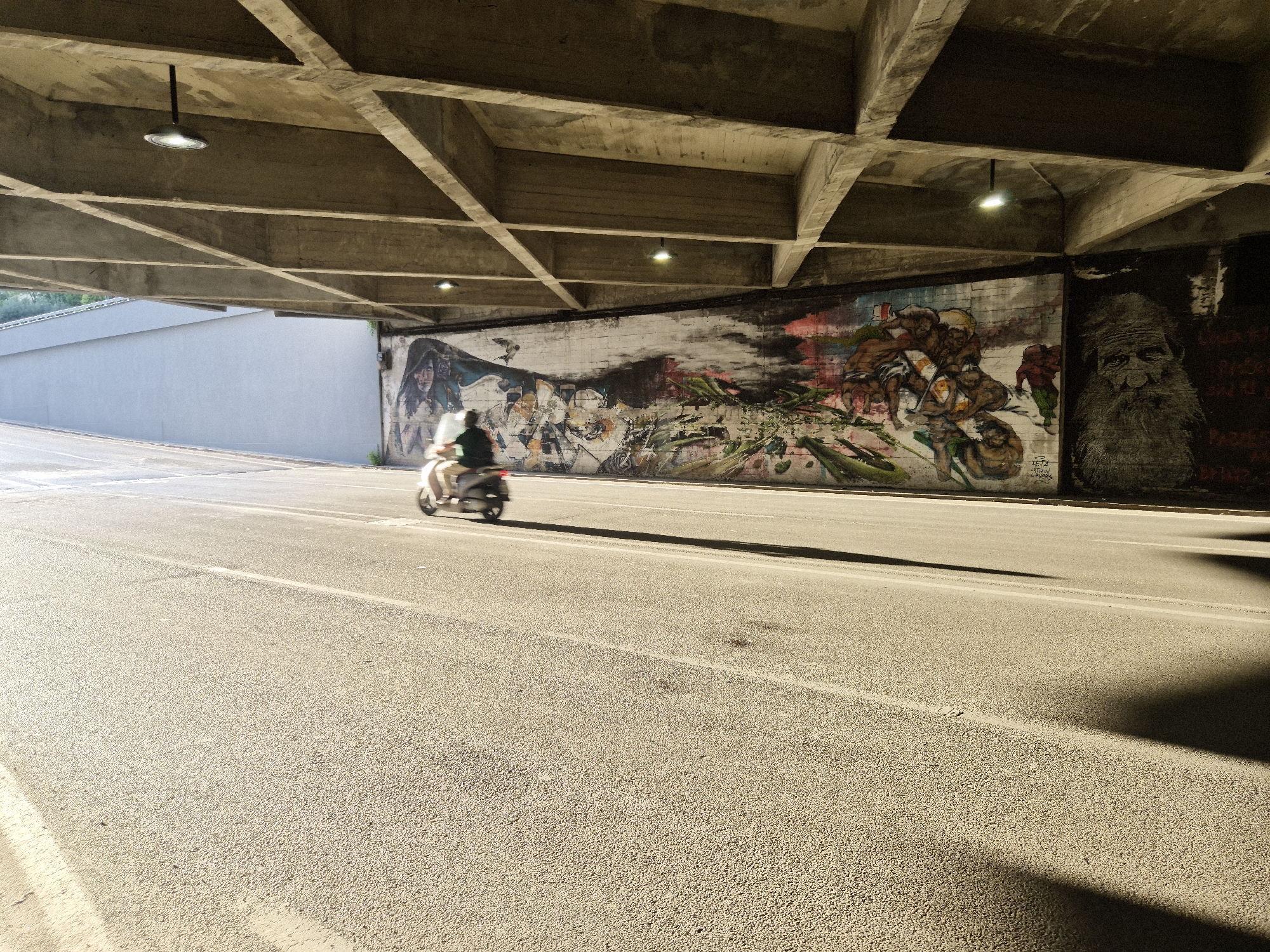 An overpass in the city of Taranto, Italy, with a scooter rider in motion on the road below. The overpass is constructed of concrete beams forming a grid pattern overhead. Two circular light fixtures are visible attached to the underside of the overpass. The road surface is asphalt, marked with white lines. The wall behind the road features graffiti art, including a portrait of an elderly man with a long beard on the right side, and abstract figures and a woman's face on the left. The scooter rider is blurred due to motion, wearing a dark shirt and light-colored pants, and is in the left lane of the road, heading away from the viewer. The sun casts long shadows across the road. The left side of the image shows a light-colored, textured wall, possibly part of the overpass structure.