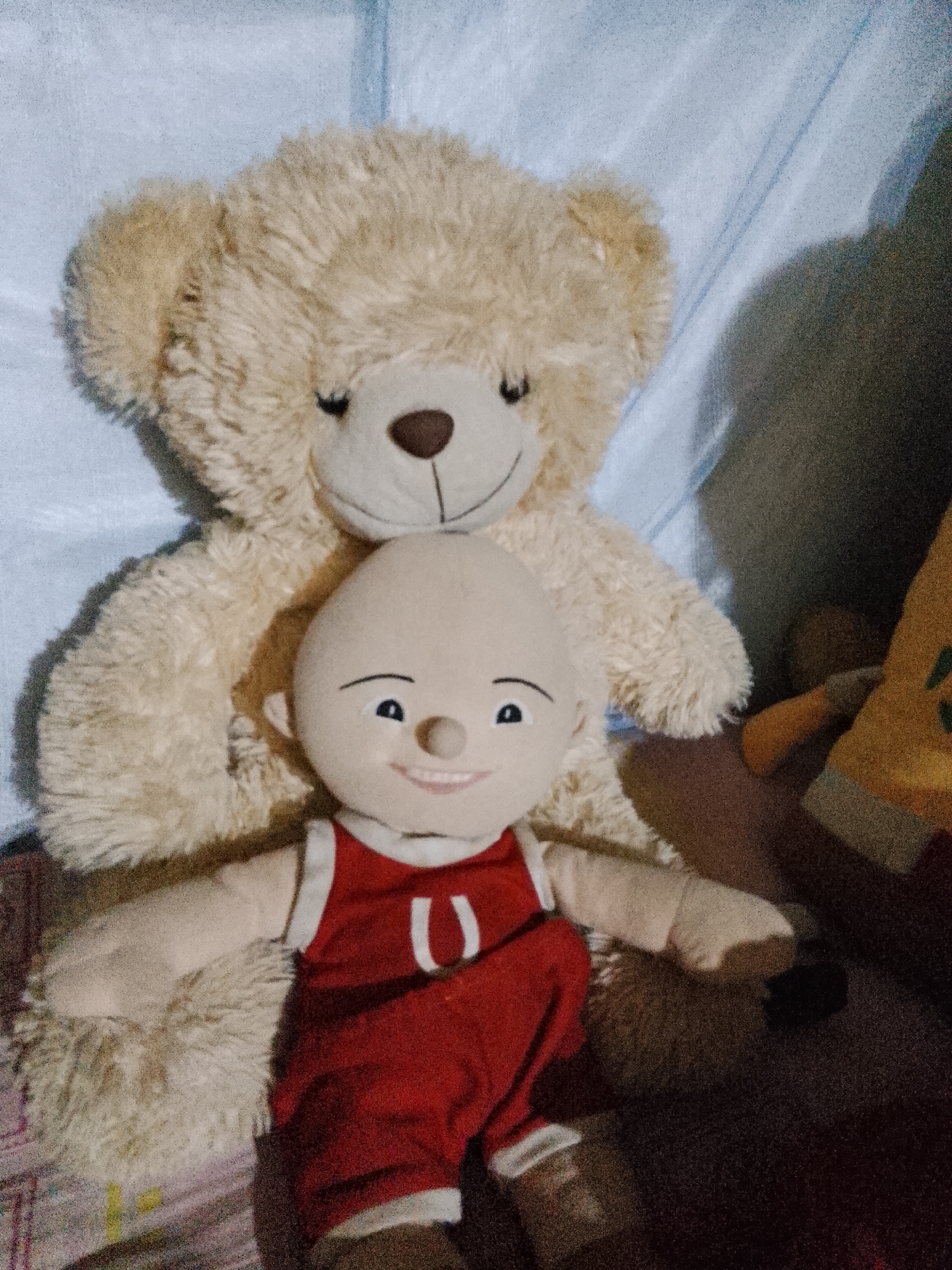 Two stuffed toys are positioned on a surface that appears to be a bed. In the foreground, a bald doll with a smiling face, black eyes, and a mouth with visible teeth is wearing a red sleeveless shirt with white trim and a white 'U' insignia. It has light-colored skin and is leaning against a larger stuffed toy. Behind the doll is a light brown, fluffy teddy bear with dark brown eyes and a dark brown nose. The teddy bear's arms are positioned around the doll. The background consists of a blue fabric, possibly a bedsheet, with a shadow cast on the right side of the image. A partially visible yellow stuffed toy with green accents is on the far right. The scene is lit in a way that creates shadows and highlights, suggesting indoor lighting conditions. No specific location in Pasuruan, Indonesia, can be determined from the image.