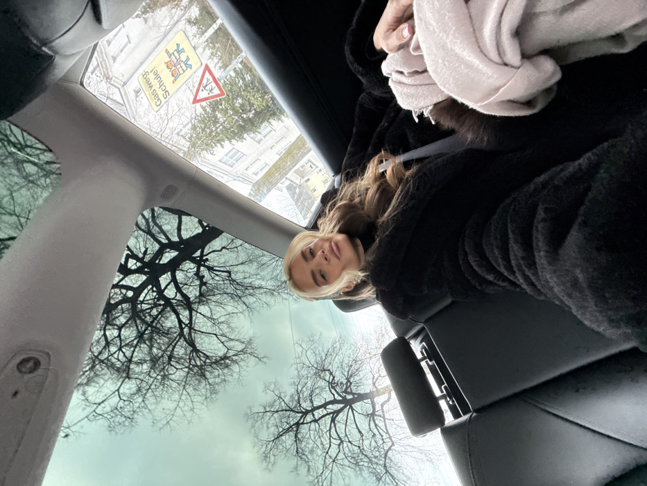 A female subject is depicted seated in the left rear passenger seat of a vehicle, looking directly towards the camera. She wears a black, textured coat over a dark turtleneck and has long, wavy hair with prominent blonde front sections. A seatbelt crosses her chest and lap. Her left hand is visible, holding a light-colored, folded garment. The vehicle's interior features black seating and a panoramic glass roof, which reflects bare tree branches against an overcast sky. Through the passenger window on the right, an urban street environment is visible, including a multi-story building and two road signs. The signs include a rectangular yellow sign with "Gas weg! Schule!" and a child graphic, positioned above a red triangular warning sign depicting two children walking. These signs confirm the location as a school zone within the city of Berlin, Germany. The bare trees indicate a cold season.