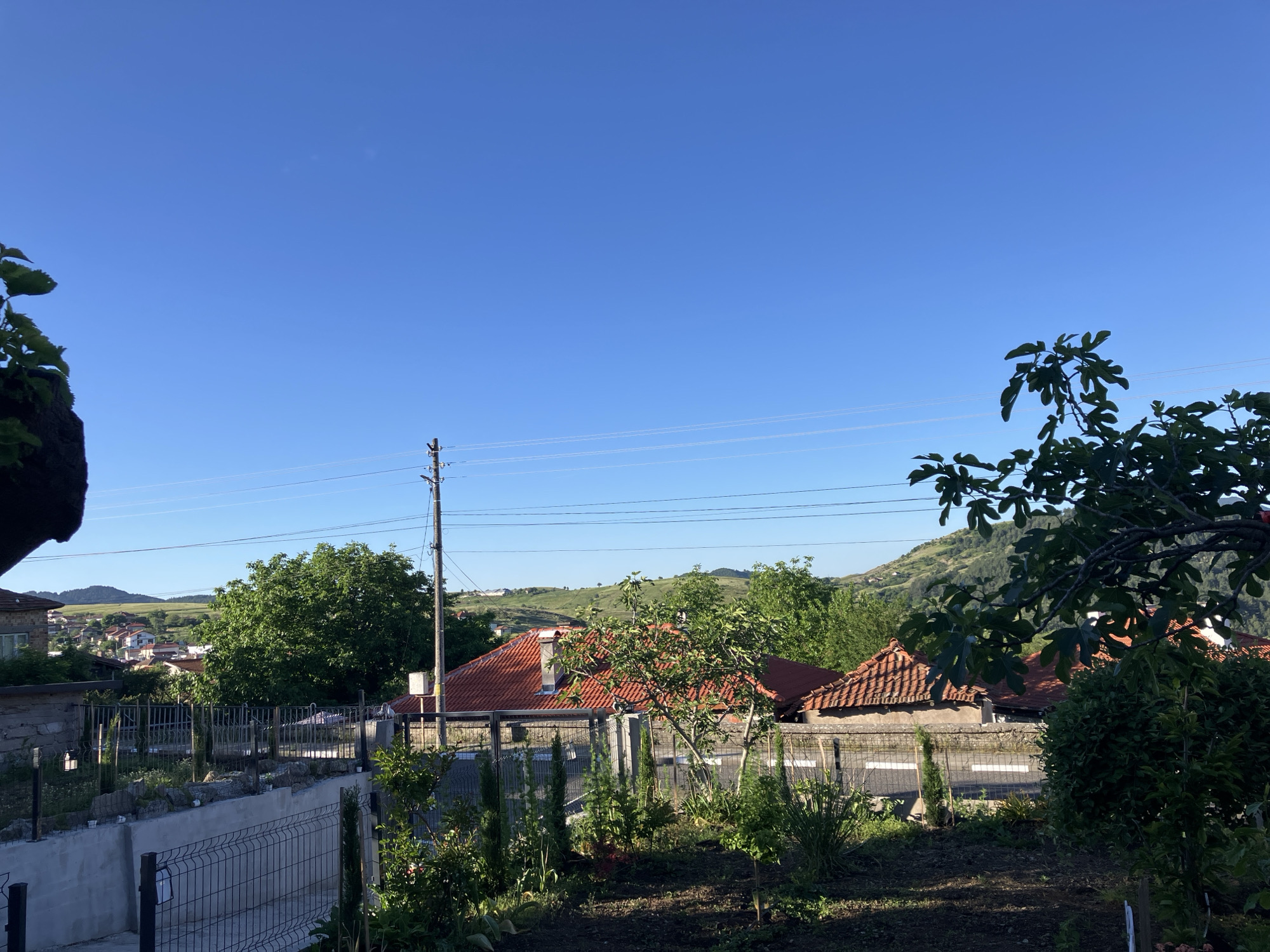 The image shows a residential area in Komuniga, Bulgaria, on a sunny day. A clear blue sky is visible above tiled roofs of houses, partially obscured by foliage from trees and bushes in the foreground. A utility pole with power lines runs across the scene. In the distance, a hillside is visible. A garden area with a wire fence and a concrete retaining wall is present.