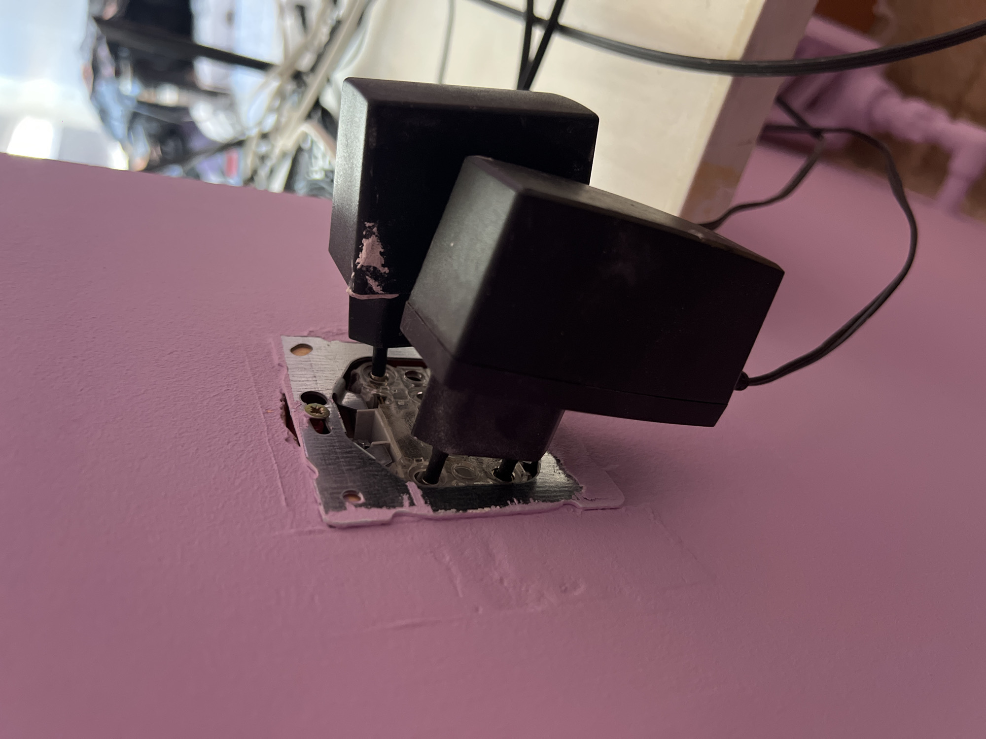 A close-up, eye-level shot captures two black electrical adapters plugged into a single wall outlet. The outlet plate, made of silver metal, is visible behind the adapters, with a screw in the upper right corner. The wall is painted a vibrant pink color, with some texture visible on its surface. The adapters themselves are simple, rectangular black boxes. One adapter is plugged in more deeply than the other, causing them to slightly overlap. The cords from the adapters extend out of the frame, suggesting they are connected to other devices. In the background, a wooden surface is visible, and further beyond that, the scene is blurred and indistinct, with hints of other objects and possibly wires. The lighting in the scene appears to be a combination of artificial light sources, contributing to the overall pink hue of the wall. The image is a still shot, capturing a moment in time without any apparent movement.