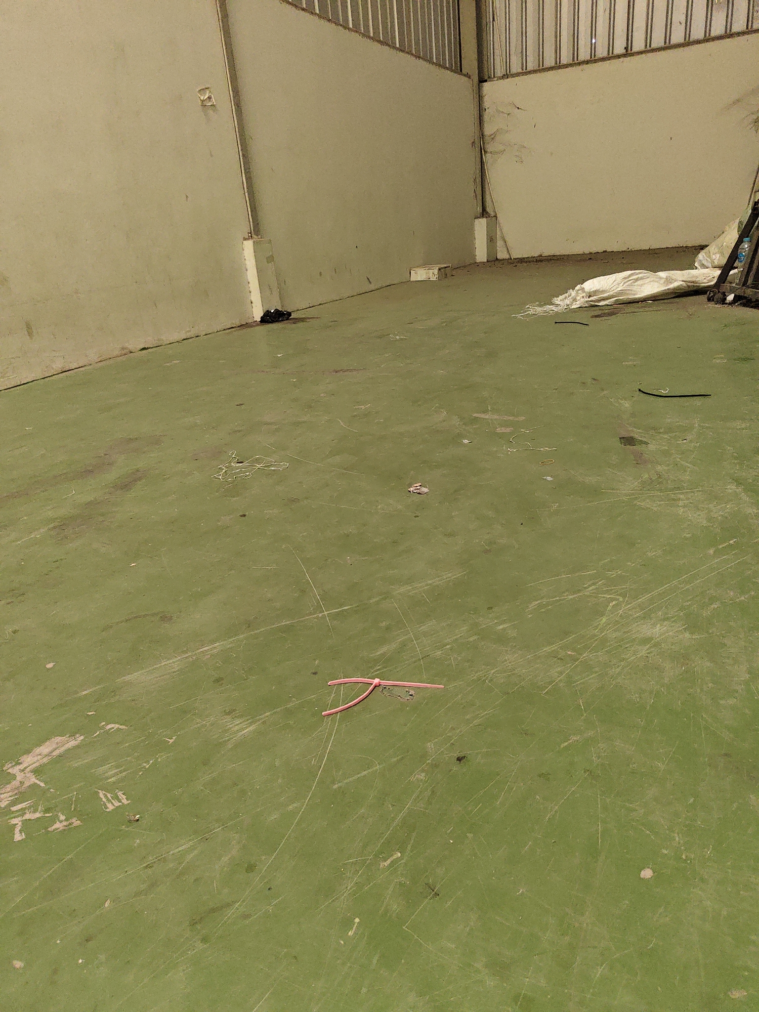 The image depicts the interior of an empty, dimly lit space, likely a warehouse or storage facility, with a light green, worn, and scratched concrete floor. The walls are a neutral, light color, and the ceiling features industrial metal grating. In the foreground, a cluster of pink plastic objects, possibly cut straws, lies on the floor. Further back, a white plastic bag is partially visible, along with a dark, cylindrical object. A small pile of dark material is present near the left wall. The overall environment suggests a disused or under-maintained industrial area. The location is identified as North Jakarta, Indonesia, though no specific landmarks or contextual elements within the image confirm this.