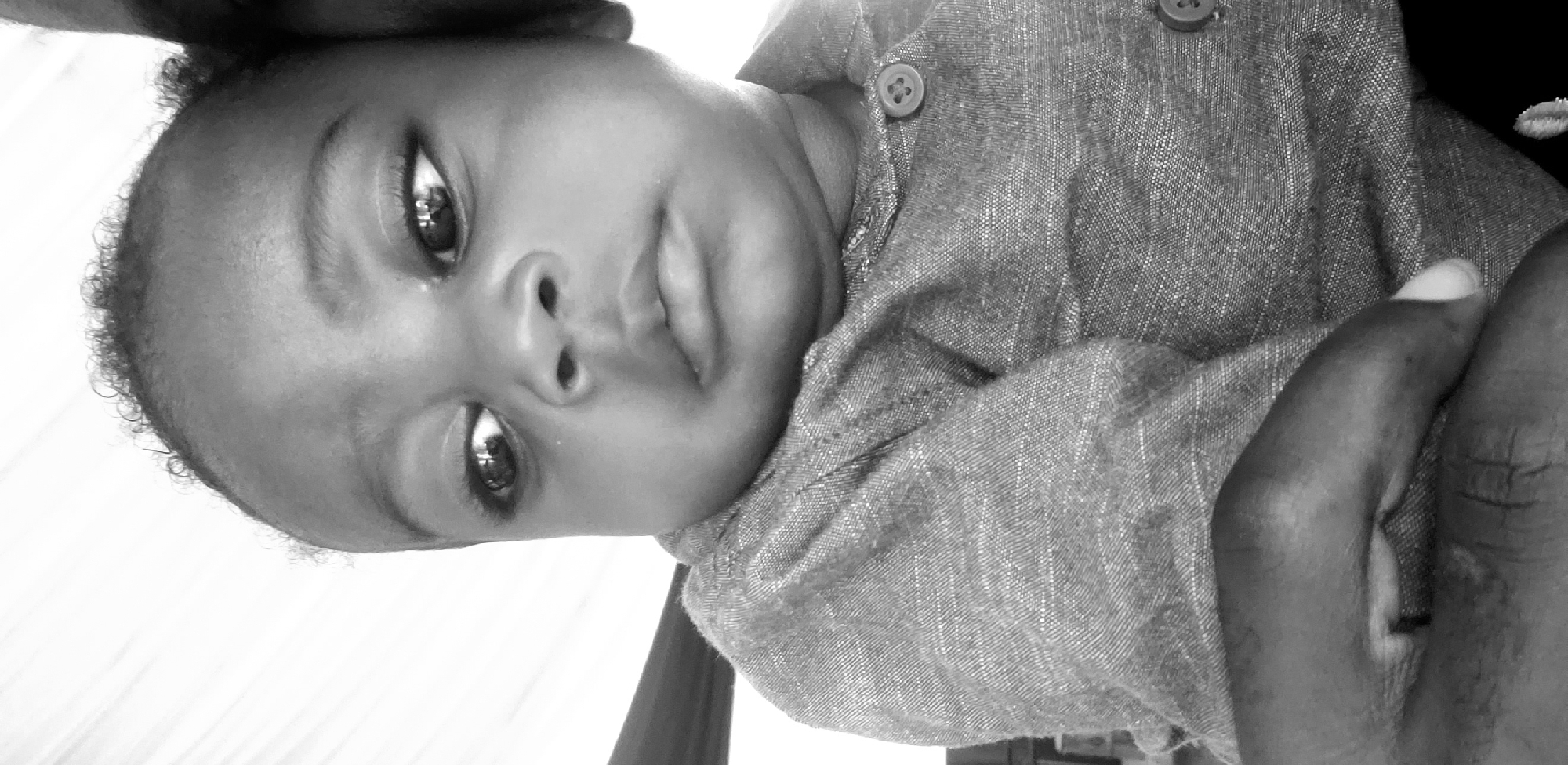The image shows a close-up, black and white, waist-up shot of a young Black child. The child is looking to the left of the frame with a neutral expression. They are wearing a gray collared shirt with two visible buttons. The background is mostly white, with some blurred dark shapes, possibly indicating a wall or furniture. The lighting suggests it is daytime. The scene is set in Narayi, Nigeria.