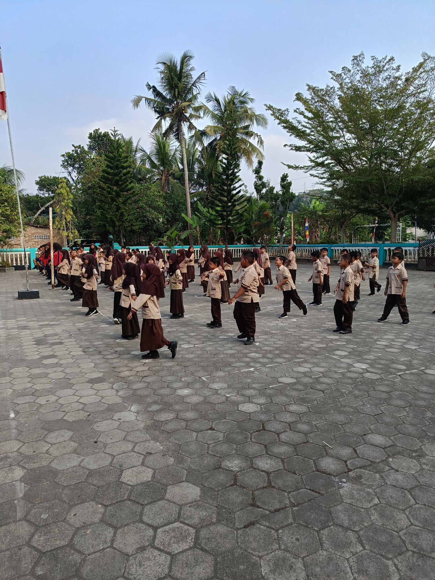 A group of students, likely elementary school-aged, in matching brown and beige uniforms are participating in an outdoor activity on a hexagonal-patterned paved area. The students are arranged in rows and appear to be performing exercises, with some engaged in synchronized movements like stretching or marching. The scene is set in an open courtyard with a low-lying building with white and blue trim visible in the background. Lush green trees, including palm trees, surround the area, and a flagpole with a red and white flag is present on the left side of the frame. The sky is mostly clear with a few scattered clouds, and the overall lighting suggests it is daytime. The location is identified as Kamulyan, Indonesia.
