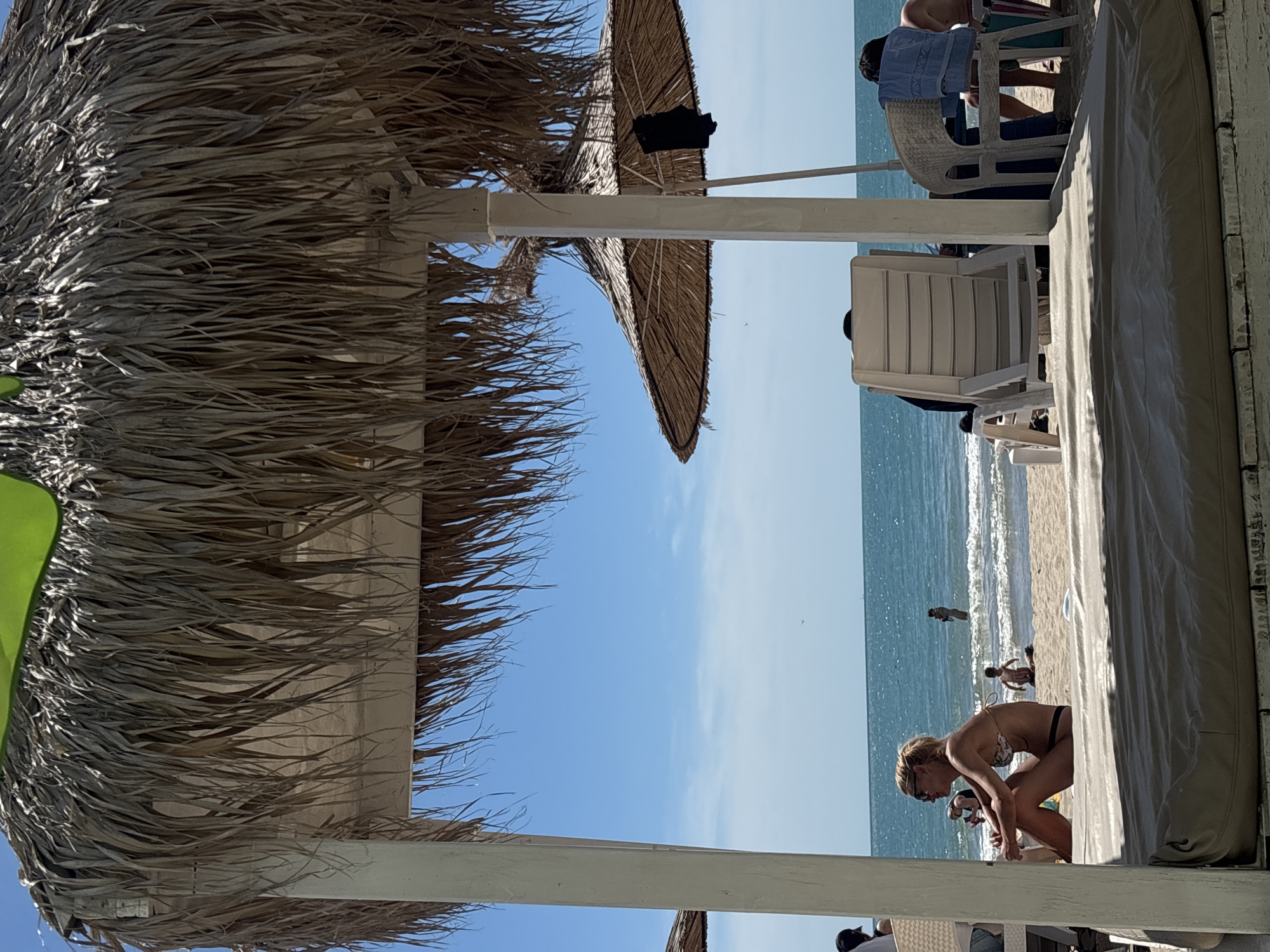 An outdoor scene at a beach, likely in Varna, Bulgaria, on a sunny day. The foreground is dominated by a thatched roof canopy with a light-colored support post in the center. Beyond this canopy, beach chairs and people are visible on the sand. In the middle ground, the turquoise sea stretches to the horizon, meeting a bright blue sky dotted with a few wispy clouds. Several people are present: a woman in a bikini and sunglasses sits on the sand in the left-center, facing left. Further back, in the shallow water, a person stands, and a child kneels on the wet sand. To the right, other beach chairs are occupied by individuals, and a towel is draped over one chair. The overall impression is one of a relaxed day at the seaside.