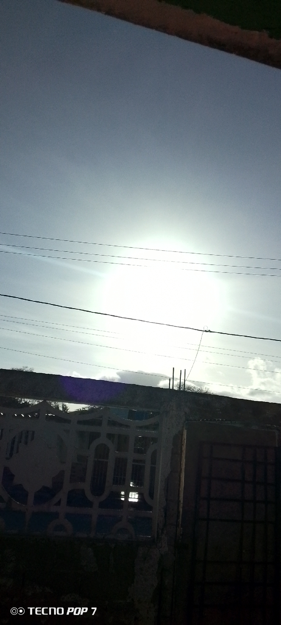 The image appears to be taken outdoors on a sunny day in Altagracia, Venezuela. The sky is bright with the sun shining through, making it difficult to see details. There are electrical wires running across the sky. In the foreground, there's a concrete structure with a decorative metal gate and a solid metal gate. The structures look somewhat weathered. Rebar sticks up out of the concrete wall. There's a hint of foliage and possibly a building visible behind the wall. The image has a "TECNO POP 7" watermark.