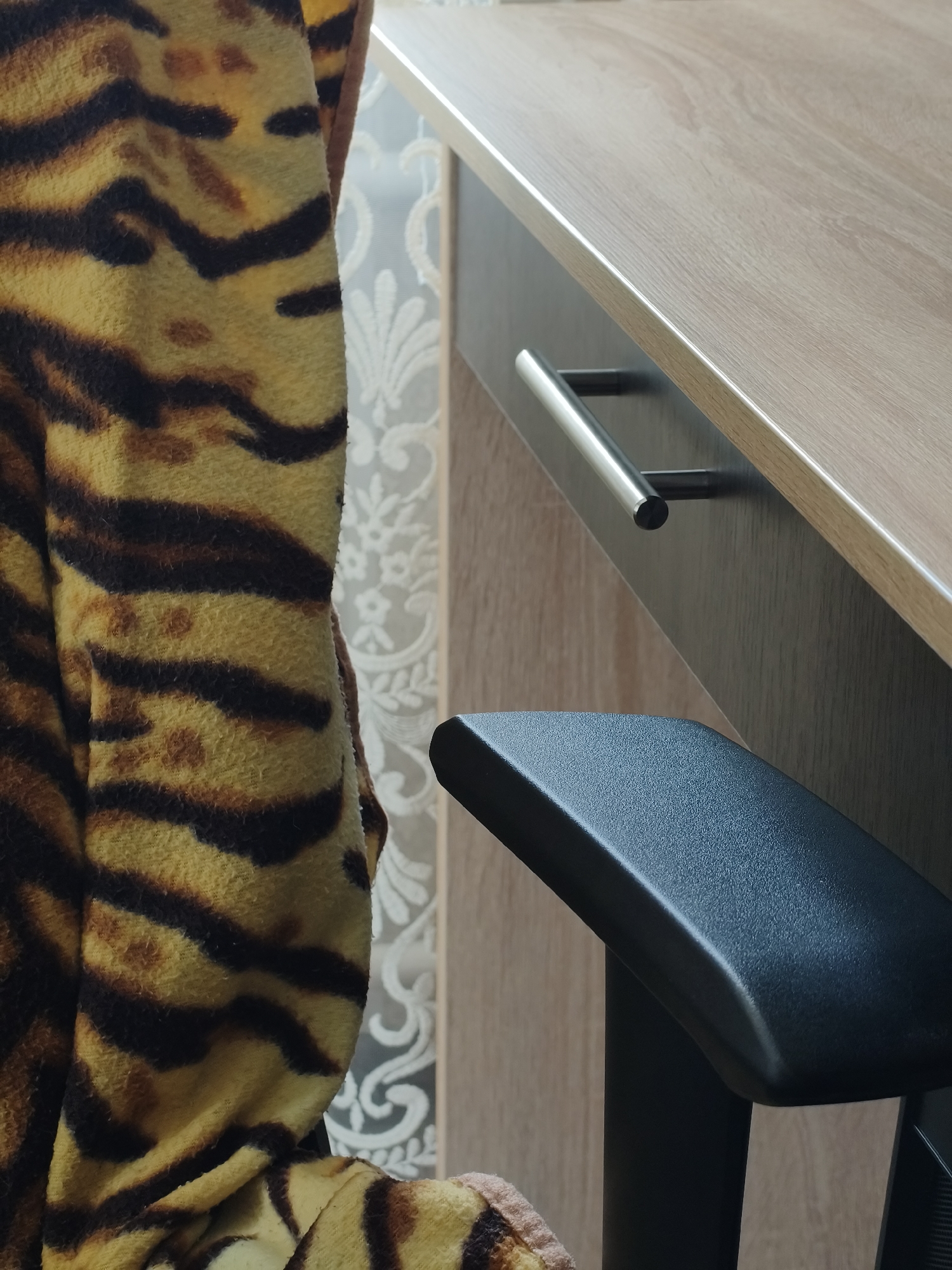 The image shows a portion of a room with an animal print (possibly tiger) garment on the left. Adjacent to it is a desk with a light wood-grain top and darker front, featuring a silver handle. To the right, the black armrest of a chair is visible. Behind the desk and garment, a patterned wallpaper in light shades is present. The location is Kryvyi Rih, Ukraine.