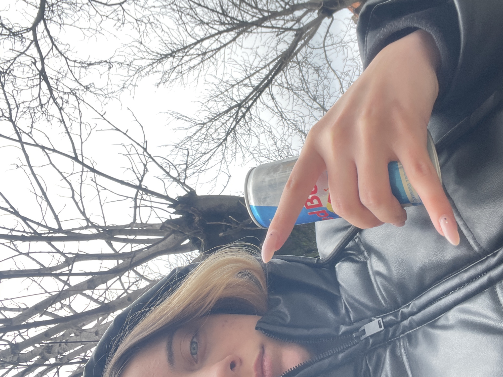 The image shows a person holding a can of Red Bull against a backdrop of bare tree branches and an overcast sky. The person is wearing a black, puffy jacket with a zipper visible. Part of their face is visible, showing blonde hair, an eye, the tip of their nose, and part of their lips. The person's hand is holding the Red Bull can. The location is likely outdoors in Sofia, Bulgaria, based on the European Red Bull can design and the local flora.