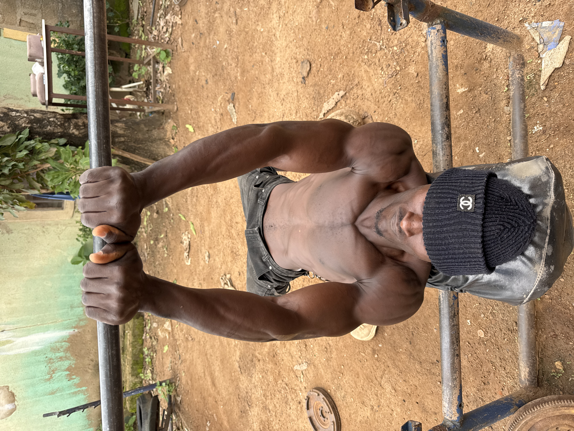 A man is performing a bench press exercise outdoors in Akwanga, Nigeria. He is lying on his back on a bench, gripping a barbell with an overhand grip. His arms are extended upwards, with his elbows slightly bent. The man is shirtless, wearing dark pants and a black beanie with a white Chanel logo. The bench is made of blue metal and appears to be old and rusted. In the background, there is a dirt ground, some greenery, and a tree. A rough wall painted green and white is visible behind the man. Weights are also present on the ground.