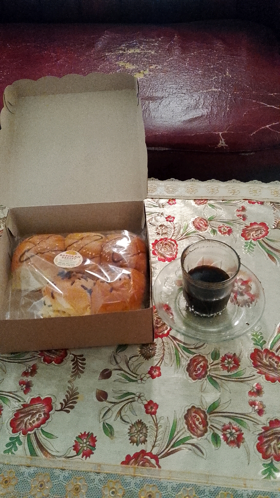 An overhead shot shows a box of pastries and a glass of coffee on a floral tablecloth. The box, which is open, contains several pastries wrapped in plastic. A label on the plastic reads "BAROKAH BAKERY". The pastries appear to be sweet rolls, some with chocolate drizzle and sesame seeds. Next to the box is a small glass of dark liquid, likely coffee, on a clear saucer. The tablecloth is light green with red and brown floral patterns and a lace border. In the background, a dark, textured surface, possibly a sofa, is visible. The scene is set in Seputih, Indonesia.
