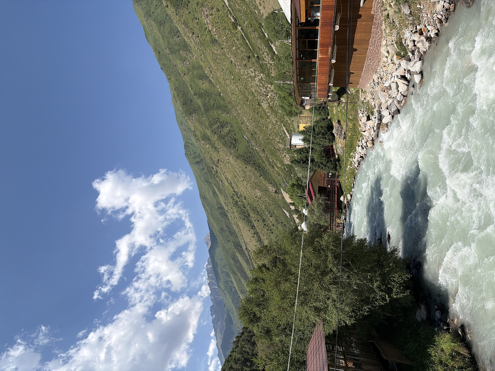 A wide, turbulent river flows through a mountainous valley under a blue sky with scattered white clouds. On the left bank, a wooden structure with a red roof is visible, partially obscured by a large green tree. On the right bank, a multi-level wooden building with large windows and a wooden balcony overlooks the river. The riverbed is lined with rocks, and the surrounding hillsides are covered in green vegetation and scattered trees. In the background, snow-capped mountains rise against the horizon.