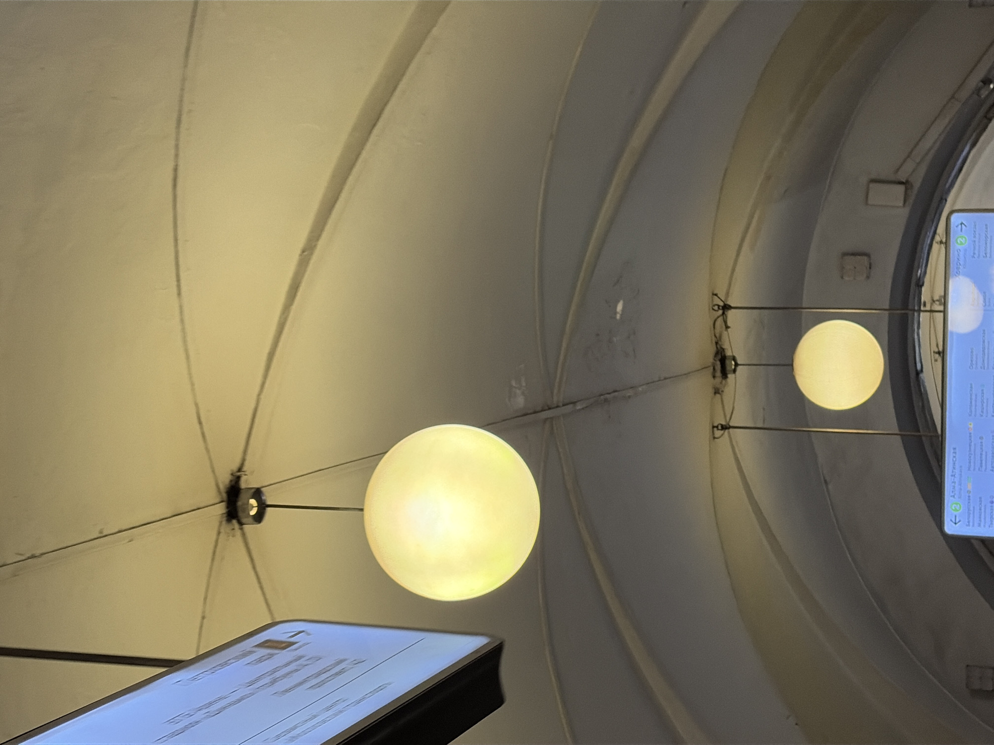 An indoor shot captures a ceiling with an arched design, possibly in a subway station. Two round, illuminated light fixtures hang from the ceiling, casting a warm glow. The larger of the two is in the foreground and is a bright, diffused yellow sphere. The smaller fixture is further back and to the right, also a yellow sphere, but dimmer and partially obscured by the ceiling's architecture. To the far left, a portion of a digital display screen is visible, showing text that is not legible. The ceiling itself appears to be a light, creamy color and has visible seams and a central junction from which the lights are suspended. The lower part of the image shows another digital display with text, also unreadable, and a portion of the arched ceiling structure. The scene is lit by the hanging fixtures, creating a soft, ambient light.