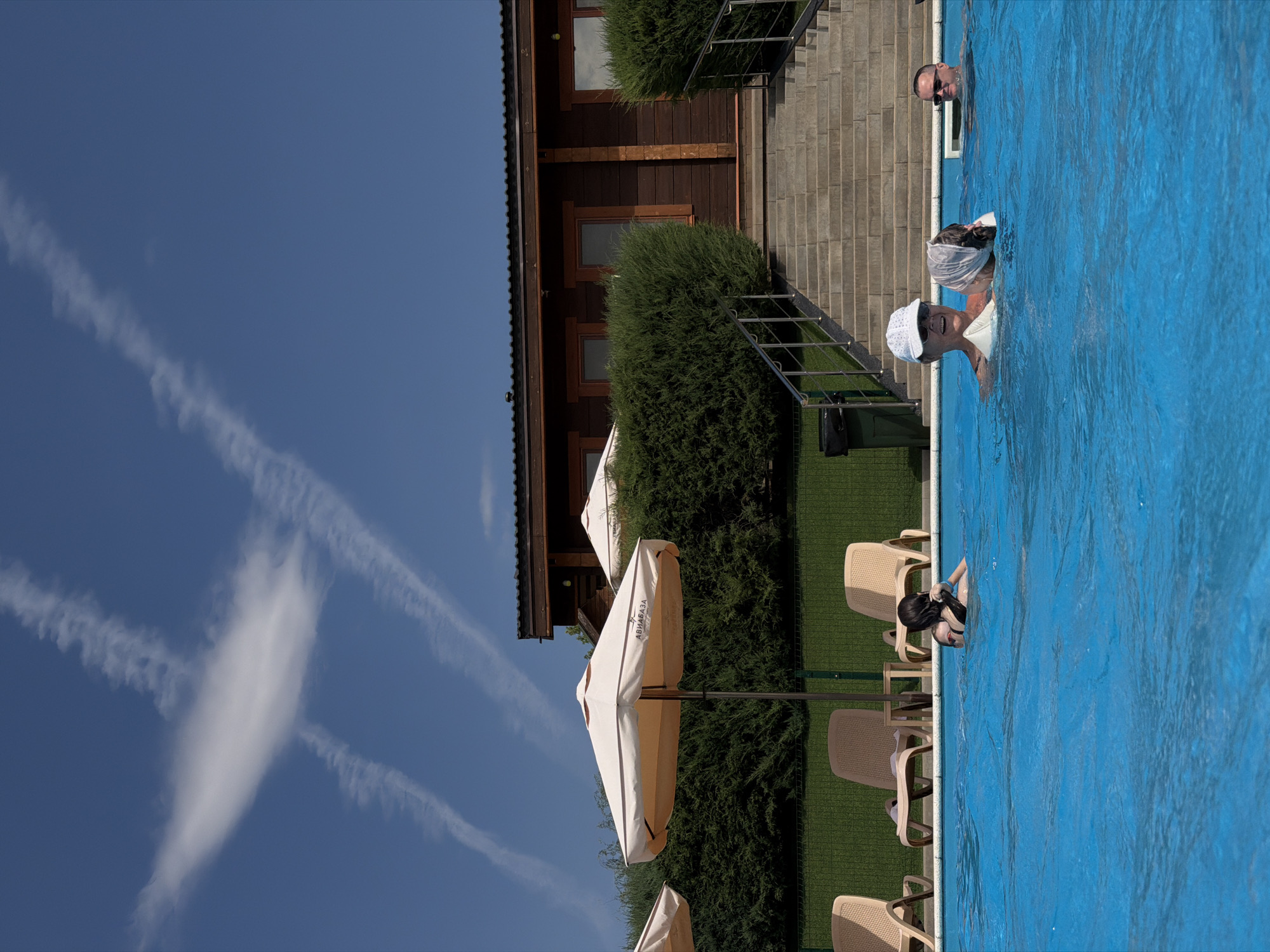This is a medium shot, taken from a low angle, of people in a swimming pool. The pool is a bright blue. In the foreground, there are three people in the water. On the left, a young person with dark hair is partially submerged, looking towards the right. In the center, an older person wearing a white cap and a white float is looking towards the camera and smiling. Next to them, a person is mostly submerged, wearing a grey head covering. On the right, a man wearing sunglasses is partially submerged, looking towards the left. In the background, there are lounge chairs and large umbrellas. Behind the pool area, there is a wooden building with stairs leading up to it. A thick, green hedge runs along the base of the building. The sky above is a clear blue with wispy white clouds. The image appears to have been taken outdoors on a sunny day. This scene is likely located at the "Aviabaza" complex in Kamyshin, Russia, as indicated by the text on one of the umbrellas.