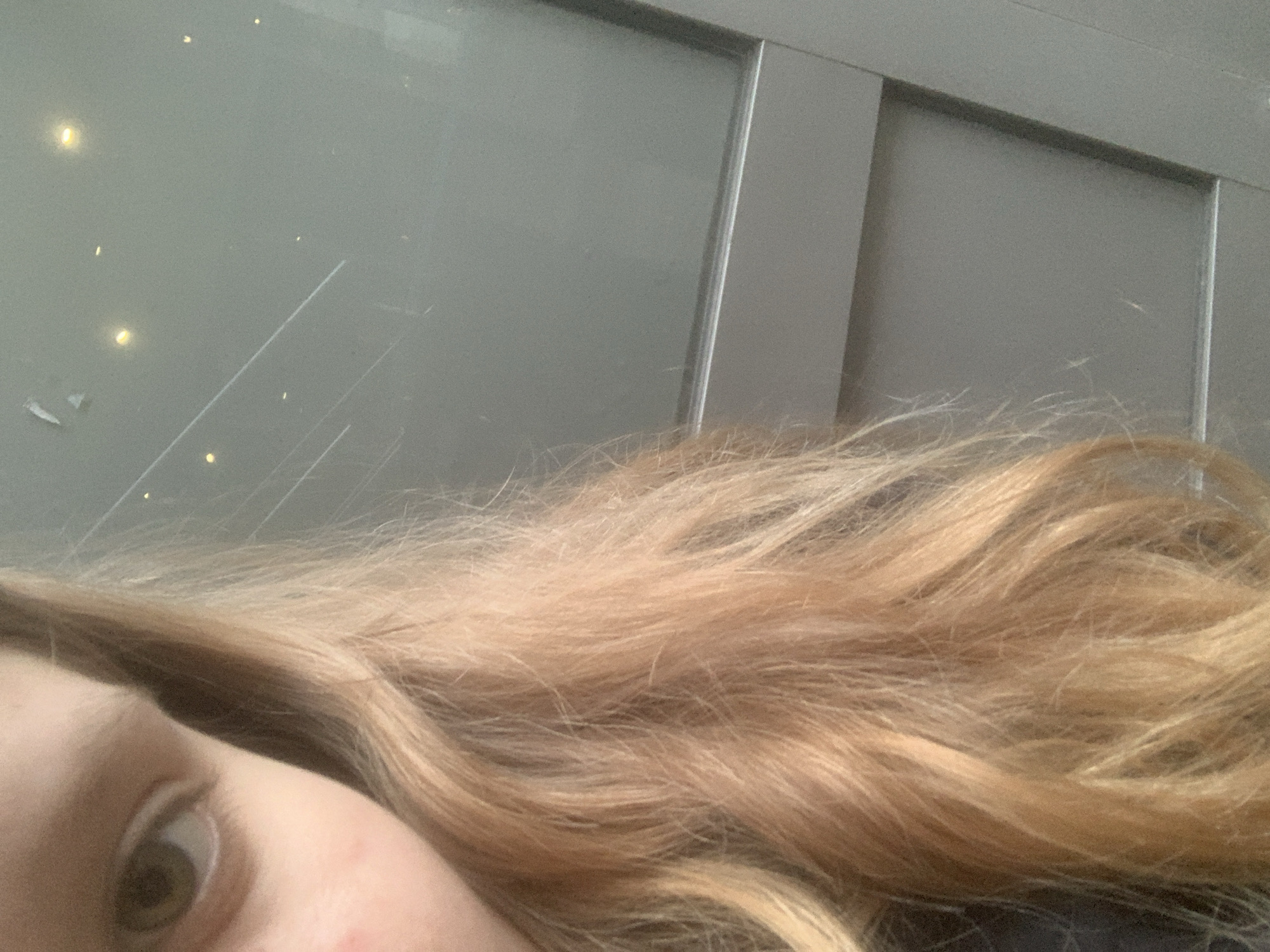 A close-up shot shows a person's face, with a visible eye and part of their skin. Their hair, light brown or blonde, fills a significant portion of the frame. In the background, out of focus, there's a textured, gray surface, possibly a wall or a large panel. The image does not contain sufficient details to determine whether the location is in the city of Saint Petersburg, Russia, or even infer it based on the limited context.