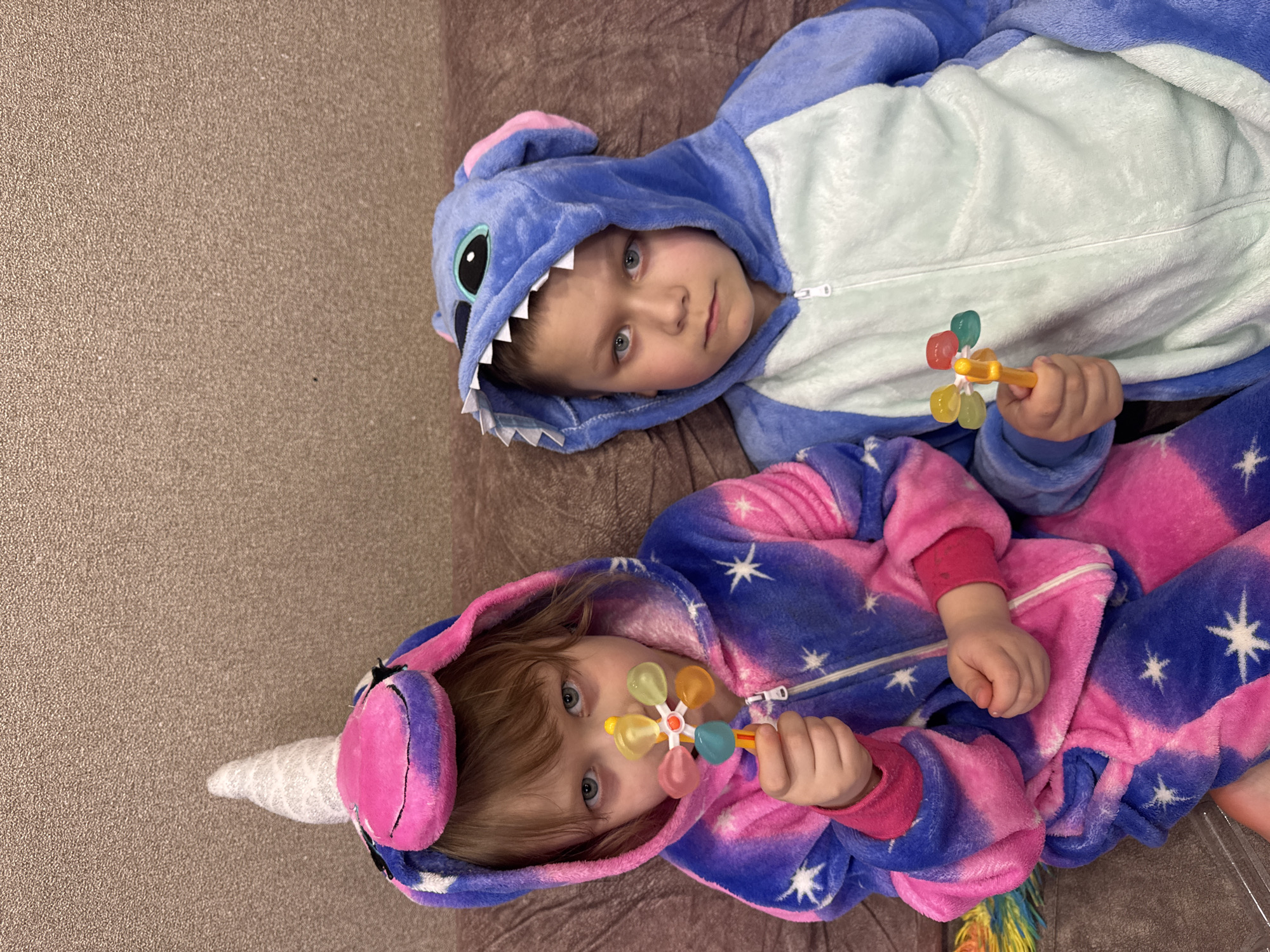 Two young children are captured lying down in an indoor setting. The child in the upper left wears a blue onesie resembling the character Stitch, complete with a hooded head featuring large eyes and jagged white teeth. This child has light blue eyes and a direct, neutral gaze towards the camera. They hold a yellow toy with multiple translucent, colorful flower-shaped components in their right hand.
The second child, positioned lower and to the right, is dressed in a vibrant pink and blue unicorn onesie patterned with white stars. This outfit includes a hooded head with a white unicorn horn and pink and purple ear details. This child also possesses light blue eyes and gazes directly at the viewer with a similar composed expression. They are holding an identical colorful, flower-shaped toy in their left hand.
Both children are resting on a textured, light brown surface, possibly a carpet or sofa. The lighting is soft and even, typical of an indoor environment. The scene depicts a calm moment, possibly during playtime or rest, with the children looking towards the person taking the photograph. The location context provided is Chaikovsky, Russia. No visible text is present in the image.
