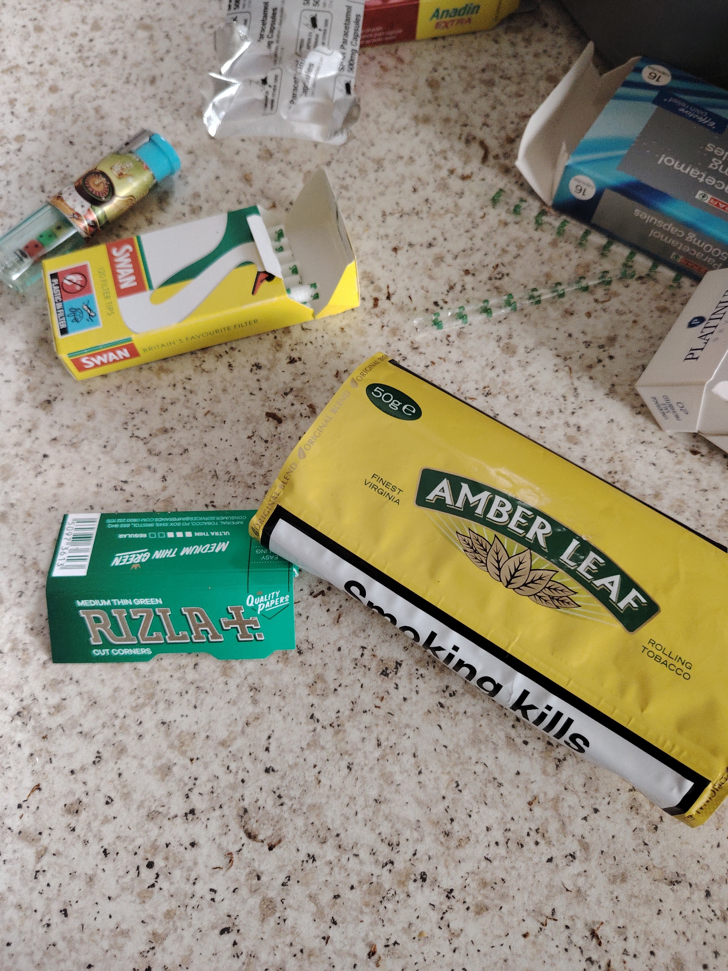 The image displays a collection of personal items and pharmaceutical products arranged on a speckled, light-colored countertop, consistent with a domestic setting in Huntingdonshire, United Kingdom.
The primary objects are:
1. **Amber Leaf rolling tobacco:** A yellow plastic pouch, marked "50g e," with "Finest Virginia" and "Rolling Tobacco" printed on it. A health warning stating "Smoking kills" is visible along the bottom edge.
2. **Rizla+ rolling papers:** A green cardboard packet, bearing the text "Rizla+" and "Medium Thin Green Cut Corners Quality Papers."
3. **Swan filter tips:** An open, yellow and white cardboard box containing "120 Filter Tips." The box is labeled "Britain's Favourite Filter" and includes an icon indicating "Plastic in Filter." White cylindrical filters are visible within the open box.
4. **Refillable lighter:** A clear blue plastic lighter, decorated with a brown and white patterned sticker or wrap.
5. **Pharmaceutical packaging:**
* Two silver foil blister packs, one partially opened, with text indicating "Paracetamol 500mg capsules."
* A blue and white cardboard box, partially visible, with "Spar Paracetamol 500mg capsules" printed on it.
* A red and yellow cardboard box, partially visible in the upper background, displaying "Anadin EXTRA."
6. **Platinum cigarette pack:** A white cardboard box, partially visible on the right, with "PLATINUM" and the number "20" suggesting a pack of 20 cigarettes.
7. **Green plastic elements:** A scattering of small, green, cylindrical plastic segments or capsules are visible on the countertop, originating from an unknown source but positioned near the Swan filter tips and Paracetamol packaging.
The items are casually arranged, suggesting recent use or placement. The countertop has minor specks and small debris. No individuals are present in the image. The branding of the products is consistent with those available in the United Kingdom.