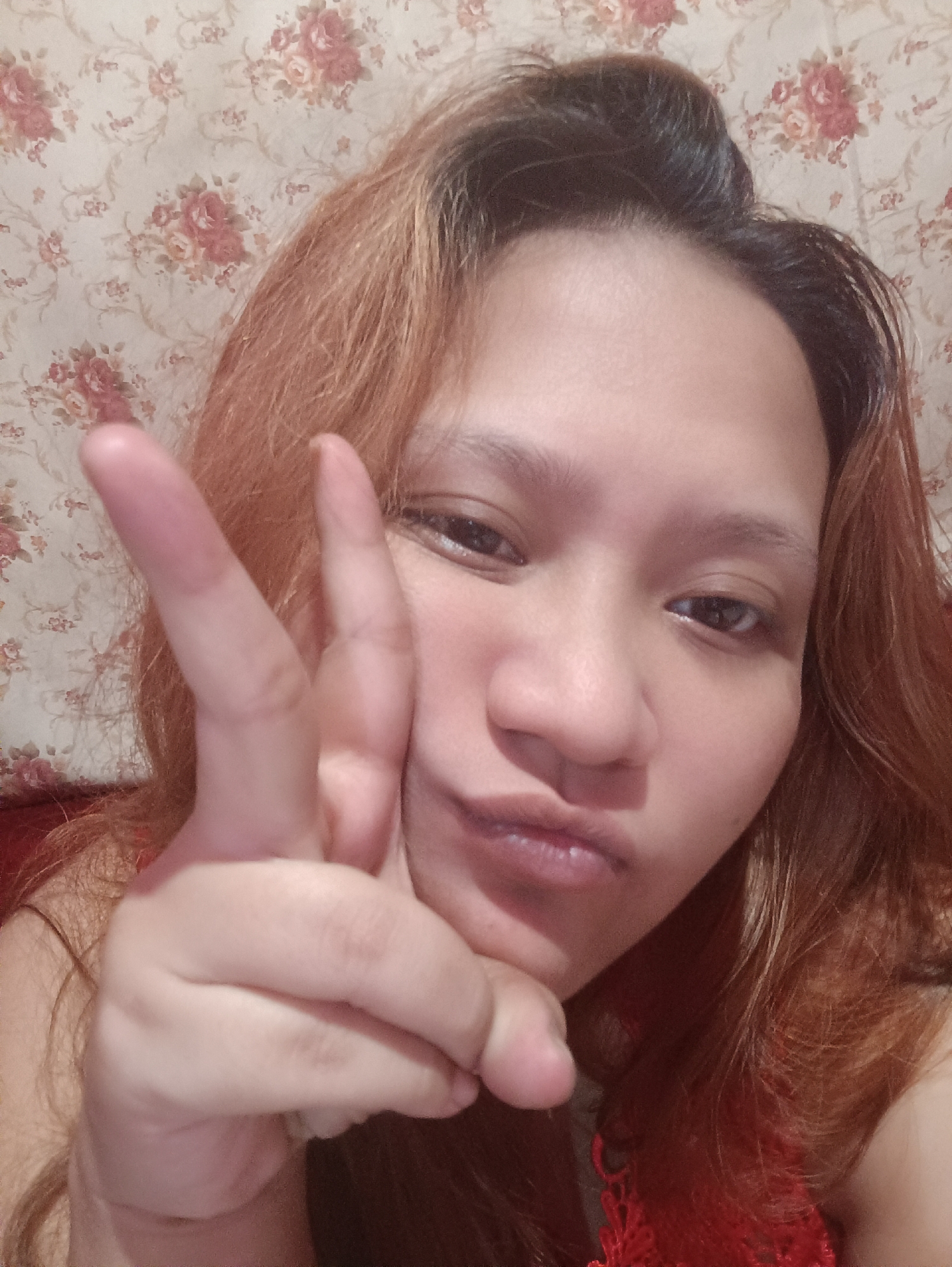 A young woman with reddish-brown hair makes a peace sign with her right hand in front of her face. She is wearing a red top. The background is a floral patterned wallpaper. The image appears to be a selfie, taken indoors. Based on visual cues such as the woman's features and the common floral wallpaper designs found in Indonesia, the location is likely Depok, Indonesia.