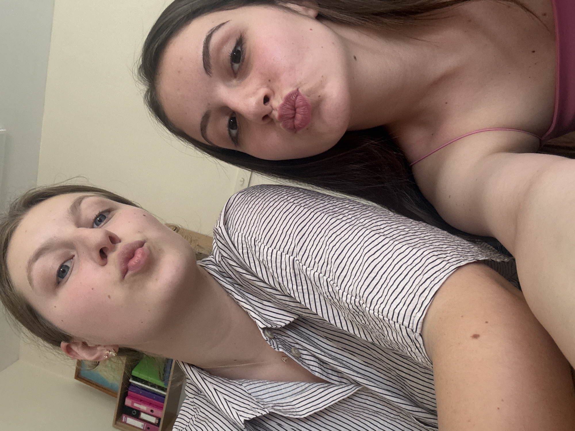 The image shows two young women taking a selfie. The woman on the left is wearing a striped shirt and has light blue eyes and blonde hair pulled back. She is puckering her lips. The woman on the right has dark brown hair and dark eyes, and is also puckering her lips. She is wearing a pink spaghetti strap top. In the background, there is a shelf with books and binders, and a framed picture on the wall. The lighting suggests an indoor setting. There is no indication of the city of Burgas, Bulgaria in the image.
