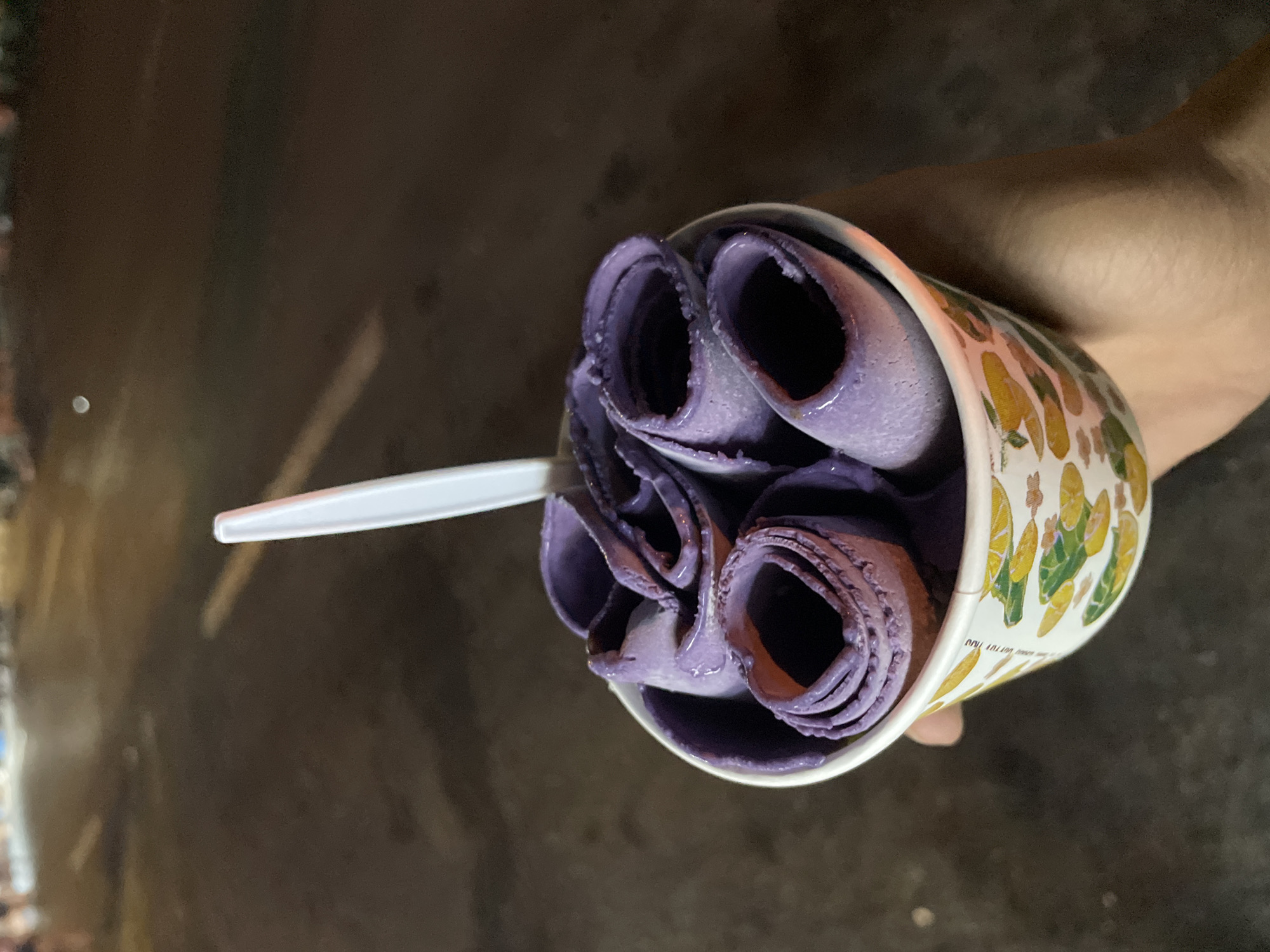A person holds a cup of rolled ice cream in Davao City, Philippines. The ice cream is purple and rolled into tight cylinders within the cup. The cup has a pattern of yellow and green fruits, possibly lemons and limes, printed on it. A white plastic spoon is inserted into the ice cream. The background is dark and appears to be a street at night, with blurred lights suggesting vehicles or street lamps. The overall scene is a close-up of the ice cream treat, with the Davao City environment hinted at in the background.