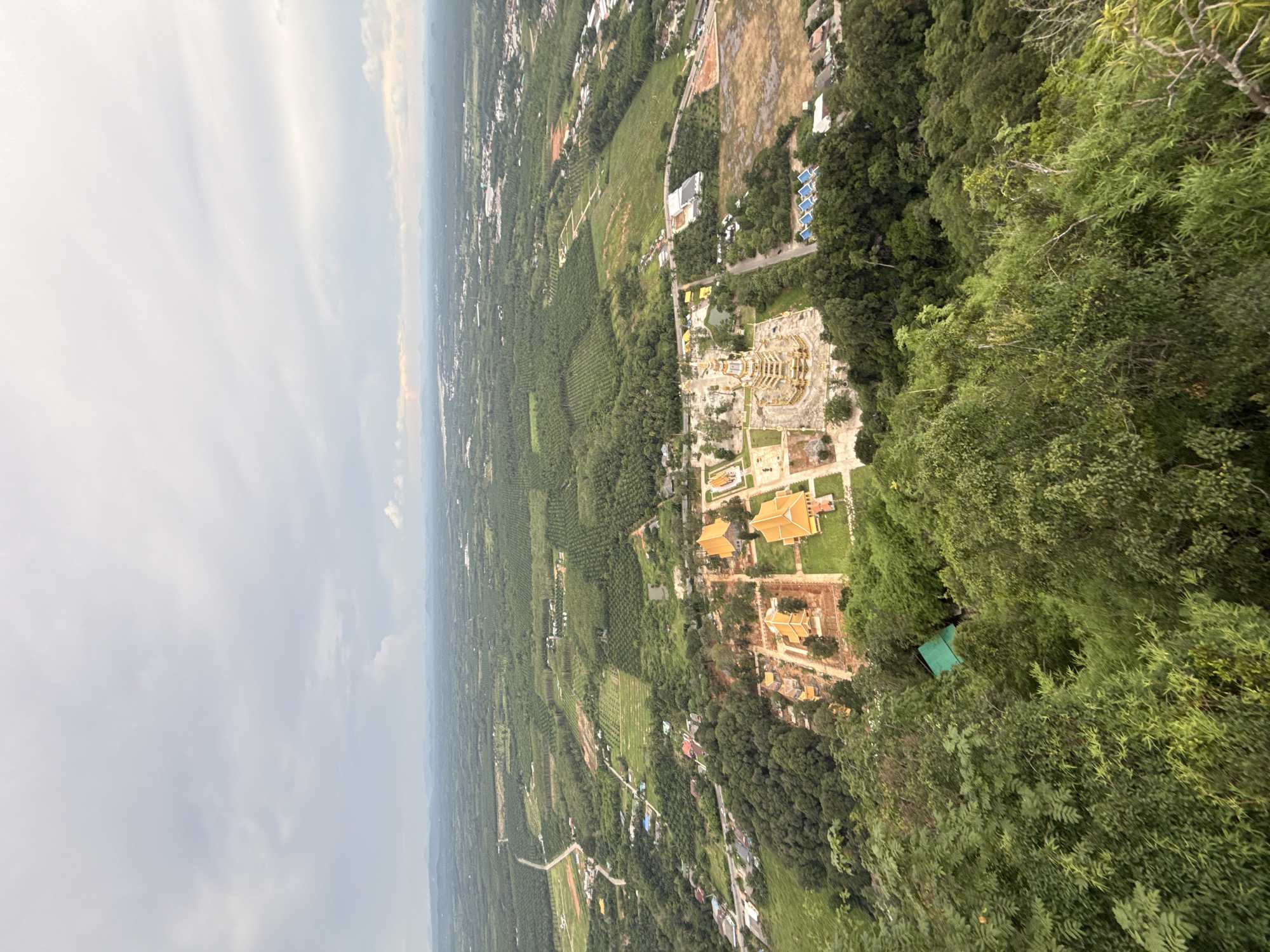 The image captures an aerial view of a lush green landscape near Ban Khao Tang, Thailand, under a cloudy sky. In the foreground, a steep, vegetated hillside descends, dotted with trees and foliage. Below, a cluster of ornate golden buildings, characteristic of a temple complex, stands out with its distinctive tiered roofs and intricate architecture. Surrounding the temple are areas of cleared land and construction, interspersed with more trees and greenery. Further in the distance, the landscape transitions into a vast expanse of cultivated fields, possibly palm oil plantations, stretching towards the horizon where rolling hills and a hazy skyline are visible. Roads wind through the terrain, connecting various settlements and agricultural areas. The overall scene depicts a rural setting with significant religious and agricultural infrastructure.