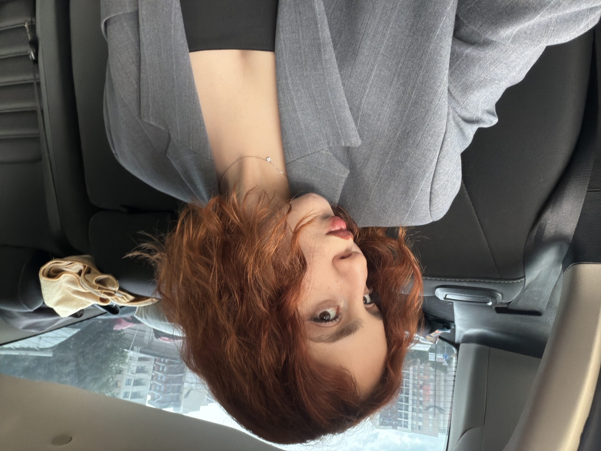 A young woman with red curly hair is positioned upside down, likely in the back seat of a car. She wears a gray blazer over a black top and a delicate necklace. The cityscape of Tbilisi, Georgia, is visible through the car window in the background, including multi-story apartment buildings. A cream-colored cloth is partially visible above the window. The interior of the car includes black leather seats and a gray seatbelt.
