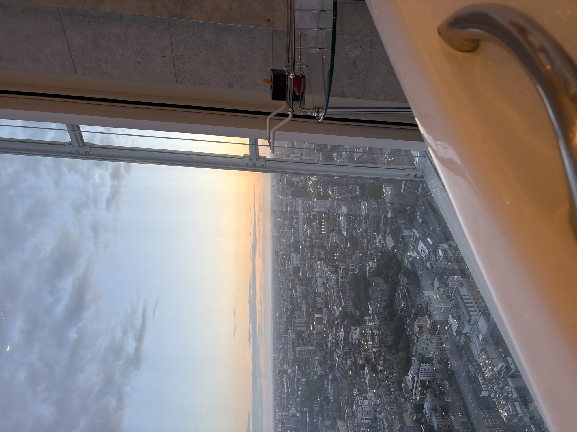 The image presents a panoramic view of a vast urban landscape from a high-rise building, looking out through a large window. The perspective suggests it was taken from an interior space, with elements of the room visible in the foreground.
On the far right, a curved, metallic handle or grab bar is partially visible, protruding from a white surface, indicative of a bathtub or shower fixture. A substantial window frame runs horizontally across the image, featuring a multi-pane design and a visible hinge mechanism with screws. Above the window frame, attached to the ceiling or upper wall, is a small black device with a red accent, possibly a sensor, control unit, or part of a window-opening mechanism.
Beyond the window, a sprawling cityscape extends to the horizon under a sky displaying the soft, warm tones of either sunrise or sunset, with gradients of orange and yellow blending into lighter blues and grays. Scattered clouds are visible in the sky. The city below is densely packed with buildings, and numerous lights within the structures are beginning to glow, underscoring the time of day as dusk or dawn. A distinctive Ferris wheel-like structure, strongly resembling the London Eye, is discernible in the distance, confirming the location as London, United Kingdom. The scene evokes a feeling of quiet observation from a considerable height. No people are visible, and no specific activity is taking place. There is no visible text.