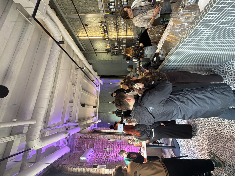 This image depicts the interior of a contemporary bar or cafe located in Belgrade, Serbia. The scene is moderately populated by individuals engaged in various activities.
**Environment:**
The venue features an industrial-chic aesthetic. The ceiling is white, exposing ventilation ducts, electrical conduits, and structural beams. Black track lighting and individual pendant lights illuminate the space. The left wall consists of exposed red brick, partially illuminated by purple ambient lighting, with a white air conditioning unit mounted high. The right wall, behind the bar, is painted a light green-grey, with a section of textured brick or tile. The floor is tiled with a repeating geometric pattern in shades of grey, black, and white.
**Objects:**
A long bar counter, faced with light blue/grey mosaic tiles, extends from the mid-ground to the far right. The bar top holds several clear glass pitchers. Behind the bar, shelves are stocked with numerous bottles of alcohol and various types of glassware. A DJ setup is visible in the mid-background-left, comprising a silver laptop and audio mixing equipment. Small, round high-top tables with chairs are situated on the far left.
**People:**
Approximately fifteen individuals are present. In the immediate foreground, a male wearing a black Helly Hansen jacket stands facing away, holding a smartphone in his right hand. To his right, a female wearing a dark top, wide-leg trousers, and sunglasses also holds a smartphone. Behind the bar, a male bartender on the far right, wearing a light grey top and jacket, is actively operating bar equipment. Another person behind the bar wears a floral top and light-wash jeans, facing away from the camera. A third individual is partially visible behind the bar, wearing shimmering gold attire. Near the DJ setup, a male with a shaved head operates the mixing equipment. Several other people are standing or seated throughout the venue, observing their surroundings or interacting with each other. A male in a brown and black jacket stands on the far left, while a female with red hair stands near a high-top table.
**Actions/Interactions:**
Patrons are primarily standing, with several individuals focused on their smartphones. The bartender is engaged in preparing drinks or maintaining the bar. The DJ is operating his equipment, suggesting music is being played. The overall atmosphere appears casual and social.