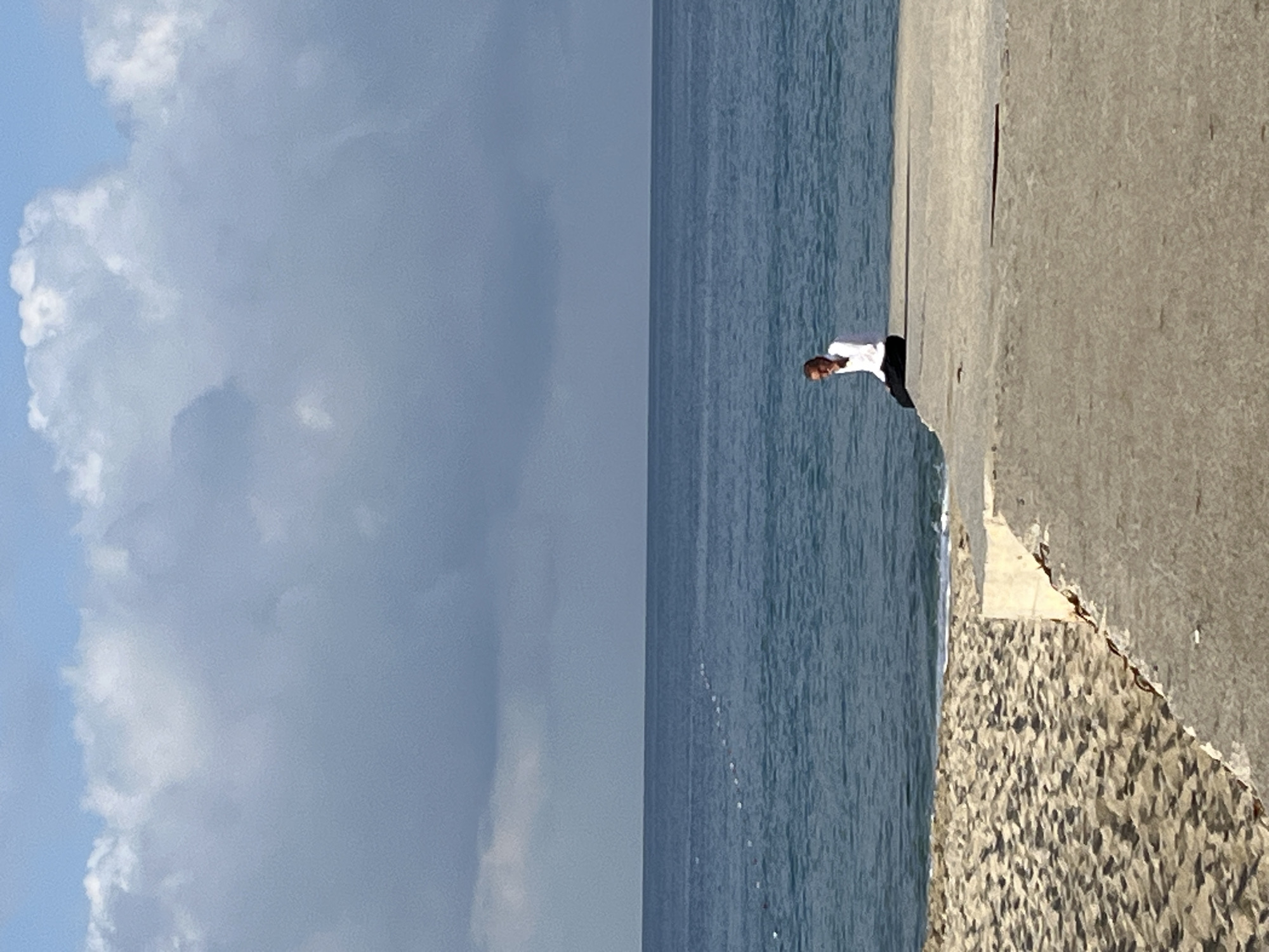 A man wearing a white shirt and dark pants stands on the edge of a concrete structure beside the sea. The structure has a rough, pebble-like base and appears to be part of a breakwater or pier. The sea is calm with a dark blue color. The sky is partly cloudy with large, billowing white clouds. The location is identified as Alanya, Turkey.