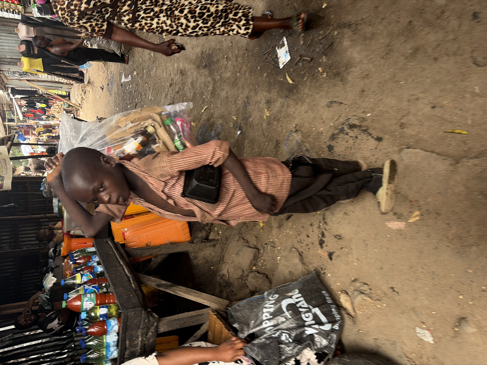 A young boy lies horizontally across a narrow, unkempt market stall in Jos, Nigeria, looking directly at the camera with a neutral expression. He wears a striped shirt (tan and reddish-brown) and dark trousers, with a small black messenger-style bag slung across his chest. His right arm is bent, supporting his head near a clear plastic bag containing small green-capped bottles and what appear to be disposable razors.
The stall he is on holds various goods, including stacked plastic bottles of liquids (some containing red palm oil, others clear), and several orange plastic containers. A white sign partially visible on the stall's edge below him shows "OKANAIY" and part of a phone number "0700/000000".
The ground to the boy's right (and below him in the image) is a dusty, uneven dirt path, littered with debris and small pieces of trash. A large black plastic bag with white text, partially reading "SHOPPING", "VIGYAN", and "SHOPPING BAGS", lies open on the ground in the lower right corner.
In the upper part of the image, the lower body of a person wearing a leopard-print dress or skirt and sandals is seen walking past from left to right, holding a smartphone in their right hand. Further into the background, on the left side of the image, are more market stalls constructed with corrugated metal sheets. Several mannequins displaying clothes (a yellow top, dark garments) are visible, along with other vendors and shoppers, indicating a busy market environment. The lighting suggests it is daytime, possibly overcast, creating soft, diffused light across the scene. The overall impression is one of everyday activity in a vibrant, albeit rustic, marketplace.