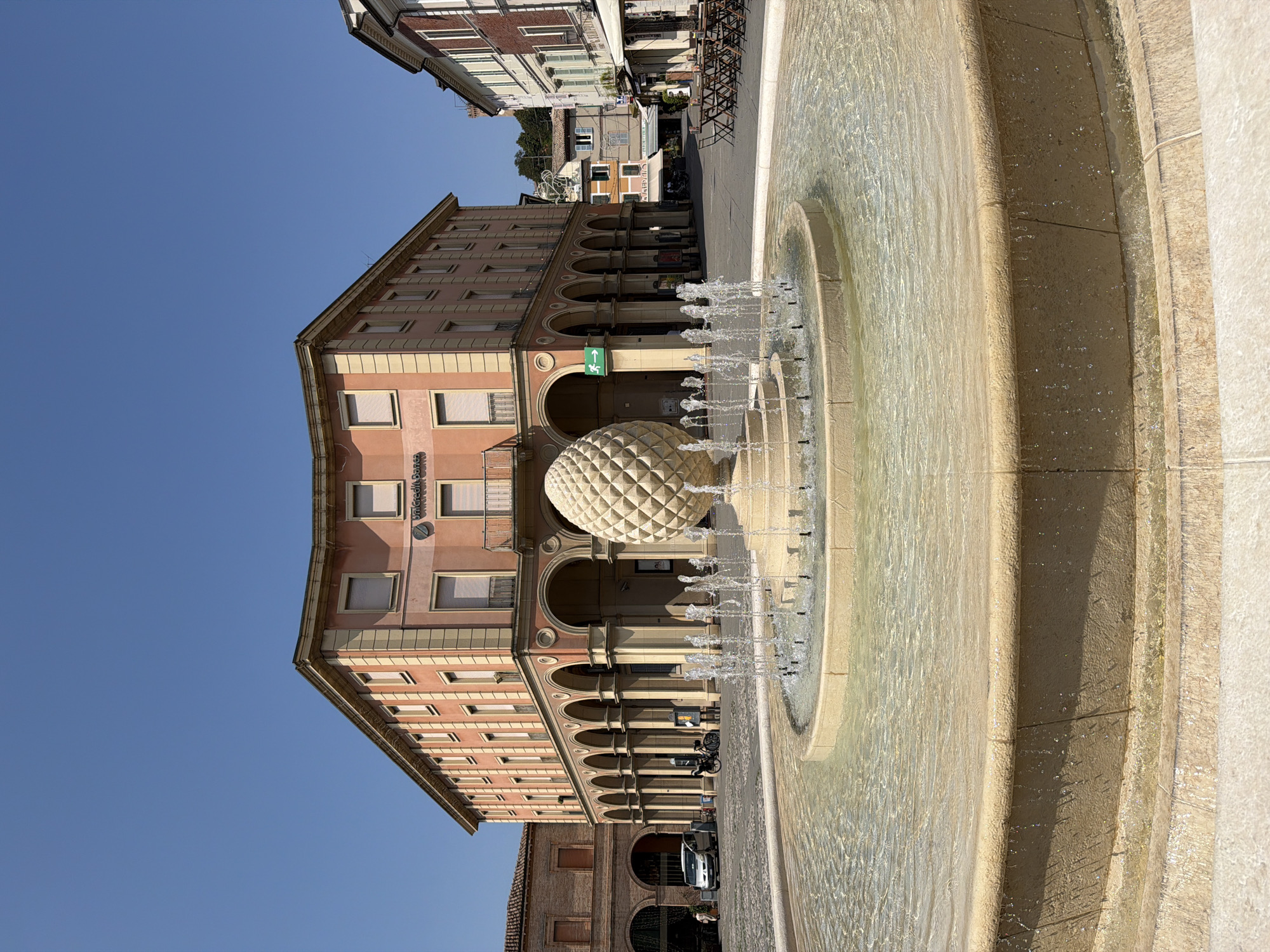 This is a full-shot, eye-level, outdoor photograph taken in the city of Santarcangelo di Romagna, Italy, on a clear, sunny day. In the foreground, a multi-tiered, round fountain with a stone basin is active, with water spraying upwards. A large, textured, light-colored sculpture resembling a pinecone or artichoke stands in the center of the fountain. Behind the fountain is a cobblestone plaza. Dominating the mid-ground is a light salmon-colored building with a curved facade, featuring a ground floor arcade with arched walkways and many windows on the upper floors. The building has an "UniCredit Banca" sign visible. To the left of this building is a brick structure with arched openings, and a silver car is parked in front of it. To the right of the salmon-colored building, other buildings line the plaza, one with red brick and arched windows, and another with a white awning and outdoor seating. The sky is a clear, vibrant blue. There are a few people visible in the scene, but they are small and not the primary focus.