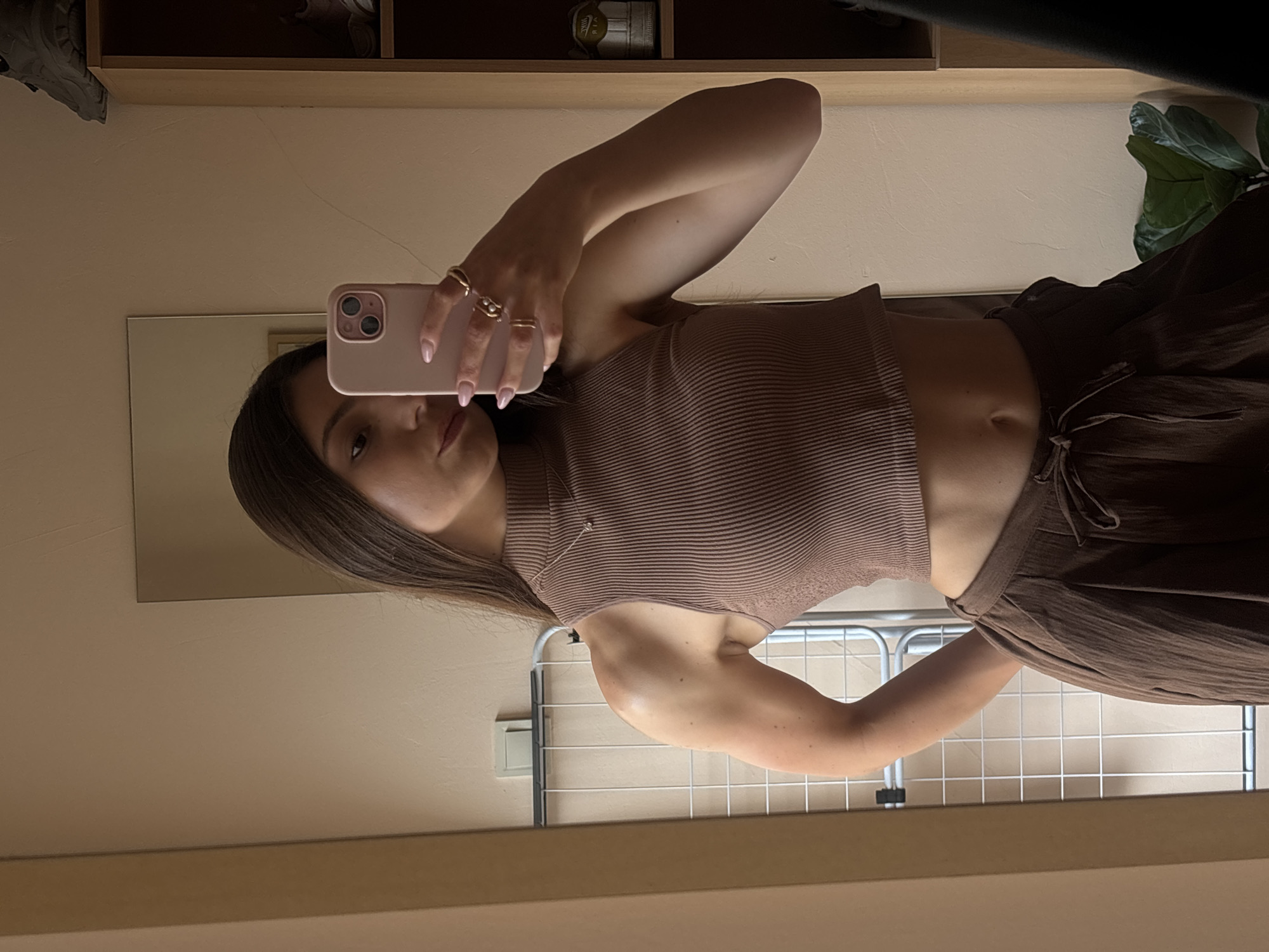 The image is a mirror selfie taken indoors by a young woman with long brown hair. She is wearing a brown ribbed sleeveless turtleneck crop top and brown wide-leg pants. She is holding a pink smartphone in her right hand, taking the photo. Her left hand is resting on her hip. She is wearing several rings on her fingers and a delicate necklace. The background shows a plain wall, a mirror, a clothing rack, and a shelf with shoes. The scene appears to be a bedroom or dressing area. There is no indication of the location being in Varna, Bulgaria.