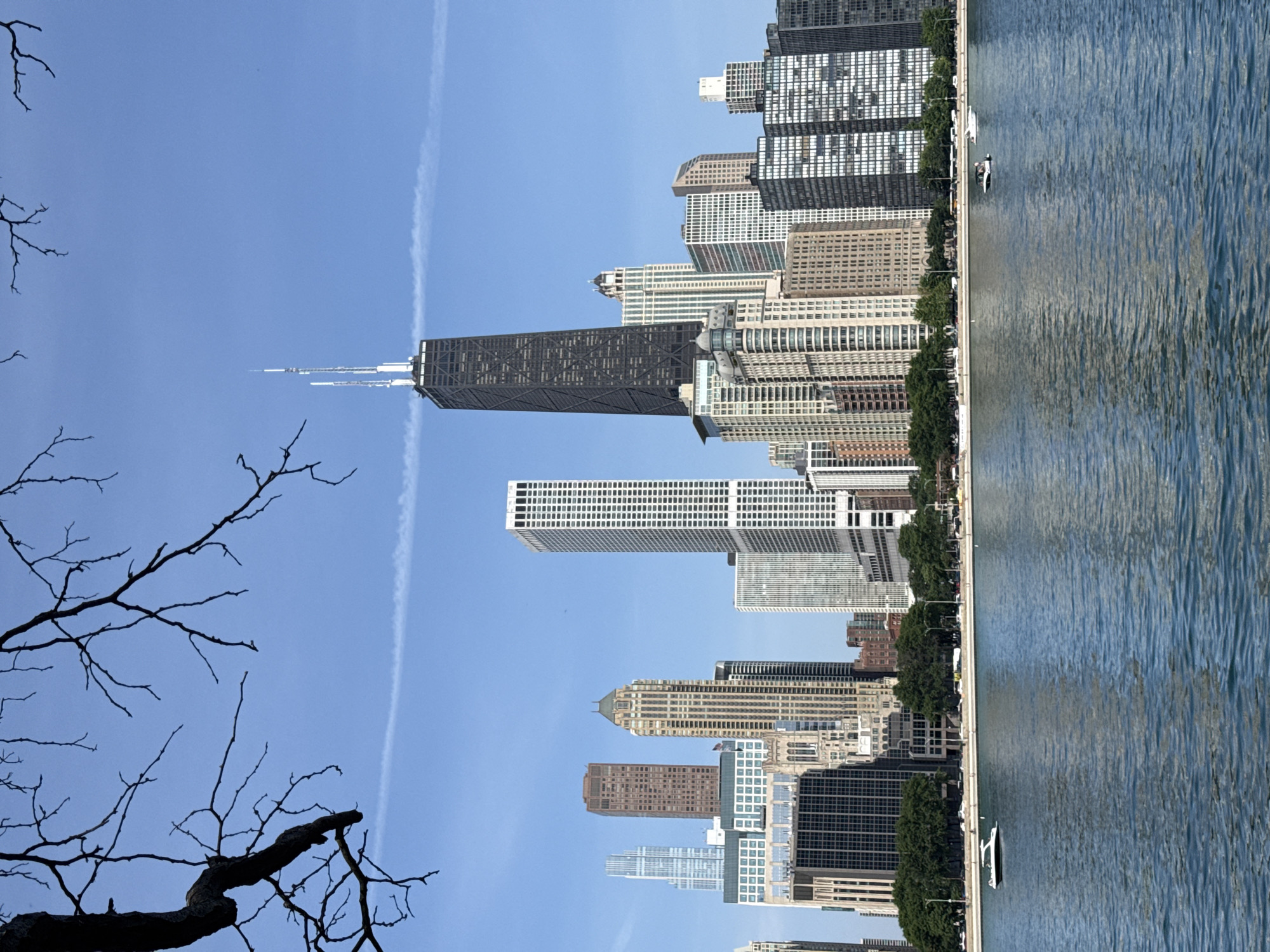 This media file captures a cityscape with a prominent skyscraper and a body of water. The scene appears to be set in Chicago, USA, as suggested by the architecture. A tall, dark skyscraper, likely the John Hancock Center, dominates the skyline, with several other modern buildings of varying heights and architectural styles clustered around it.
In the foreground, silhouetted bare tree branches are visible against a clear blue sky, indicating a cool or perhaps early spring/late autumn day. A faint white contrail stretches across the sky. The bottom right portion of the image shows a large expanse of blue water, reflecting the sky and buildings. A white boat is visible on the water, and a shoreline with trees and a sidewalk separates the water from the buildings.
The image is oriented vertically, with the sky and tree branches at the top, and the water and shoreline at the bottom. The buildings are arrayed along the water's edge. The overall impression is one of a serene urban landscape on a bright, clear day. There is no visible text in the image. The scene likely represents a daytime view, possibly late morning or afternoon, given the quality of light.