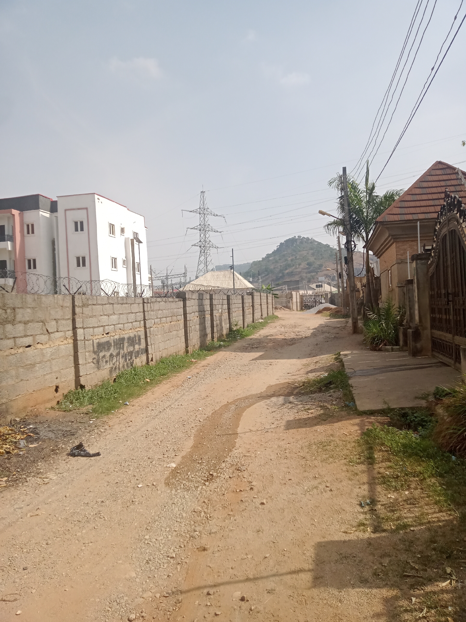 The image displays an unpaved, dusty road in what appears to be a residential or mixed-use area, likely in Gwarinpa, Nigeria, under a clear, bright sky. The road, showing tire tracks and scattered gravel, stretches into the background where it appears to curve towards a prominent rocky hill.
On the left side of the road, a long concrete block wall topped with several strands of barbed wire runs along the edge. Behind this wall, a modern, multi-story white building with red trim and numerous windows is visible. Further back, a large metal power transmission tower stands, suggesting industrial or infrastructural presence. Another building with a light-colored roof is partially visible near the base of the power tower. Faint, dark, and illegible markings or graffiti are present on the concrete wall in the mid-ground.
The right side of the scene features a narrow concrete sidewalk alongside the dirt road, bordered by patches of green grass and other foliage. A building with a distinct brown-tiled roof and an ornate, dark metal gate is prominent on this side. Overhead, multiple power lines crisscross the frame, connected to utility poles, one of which supports a street light. Palm trees and other tropical-looking plants grow along this side.
The background is dominated by a large, rocky hill or small mountain under the bright, hazy sky. No people are visible in the scene. The clear conditions and visible shadows on the ground indicate it is daytime, likely late morning to early afternoon. A small, dark, unidentifiable piece of litter is visible on the dirt road in the foreground.