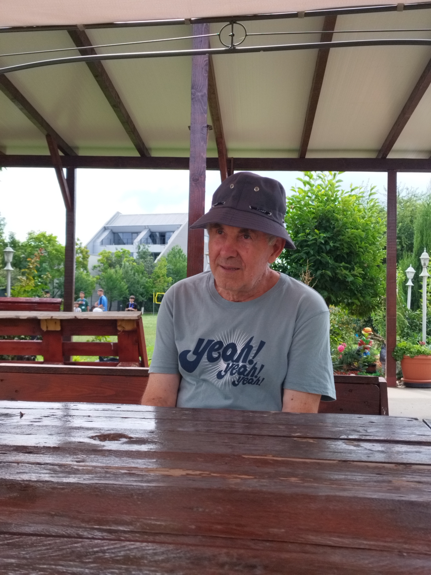 An elderly Caucasian man wearing a grey t-shirt with "Yeah! Yeah! Yeah!" printed on it in blue and a dark grey bucket hat sits at a wooden table under a white canopy. The man is facing to his left, looking slightly down. In the background, there are trees, a modern building, and other people who are out of focus. The image appears to have been taken outdoors. The scene does not provide any specific indicators of the city of Bucharest, Romania.