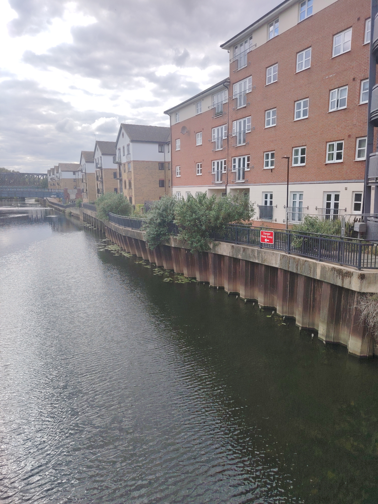 The image presents an elevated view of a wide, calm river flowing through an urban environment under an overcast sky. The sky is dominated by dense, grey and white clouds, indicating a cloudy day.
On the right side of the image, the riverbank is structured by a retaining wall composed of a concrete cap and vertical corrugated sheet piles that extend into the water. Above this wall, a dark metal fence or railing runs horizontally. Attached to this railing, approximately in the center-right, is a red rectangular sign with white text that reads "Danger Keep Away" and includes a left-pointing arrow. Behind the railing, a series of modern, multi-story residential apartment buildings lines the river. The closest building on the right features a reddish-brown brick facade with numerous white-framed windows and several balconies with metal railings, topped by a lighter, possibly cream-colored rendered upper section and a brown-tiled pitched roof. Further down the river, subsequent buildings exhibit a similar contemporary architectural style, generally featuring brick lower sections and lighter upper stories with pitched roofs. Various bushes and small trees are interspersed along the embankment between the railing and the buildings, with some bare branches visible on the far right.
The river's surface displays ripples, reflecting the grey light from the sky, and appears dark, hinting at depth or sediment. Patches of aquatic vegetation, resembling lily pads, are visible near the base of the retaining wall in the water.
In the distance, spanning the river, a prominent dark metal truss bridge, typical of a railway bridge, is visible, with a smaller, lower bridge partially obscured beneath it. No people or active interactions are visible in the scene.
This setting, characterized by modern riverside residential buildings, a waterway, and a distinctive railway bridge, is consistent with areas along the River Nene in the city of Peterborough, United Kingdom, particularly those that have undergone recent urban development or regeneration.