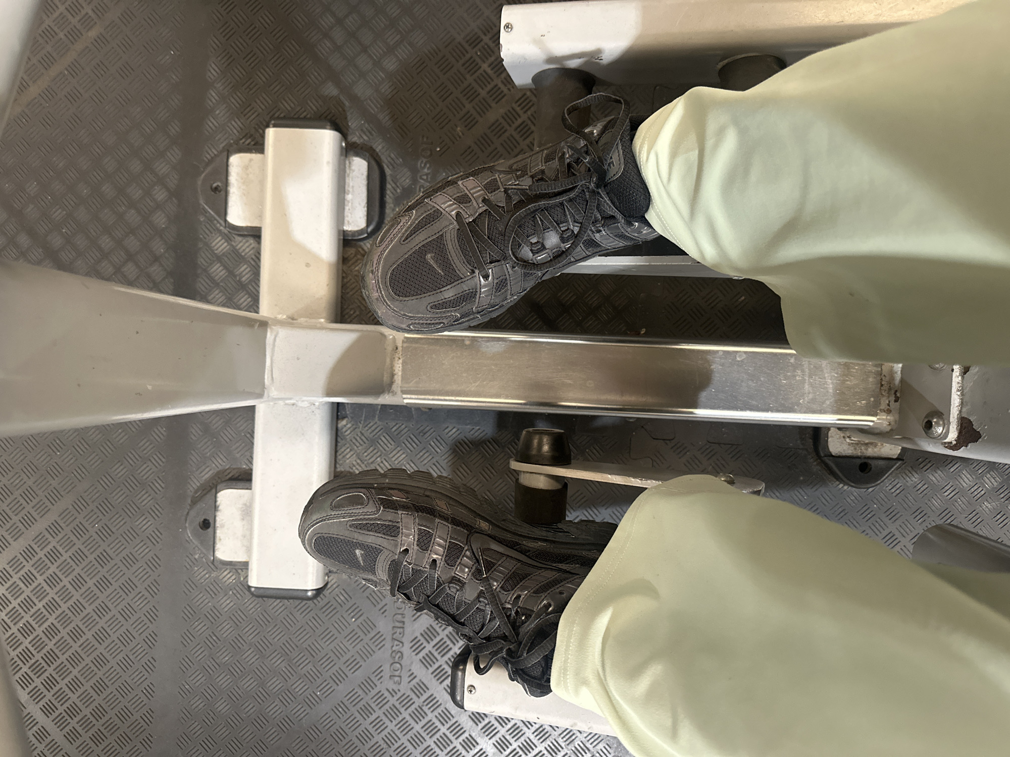 The image shows a person’s feet resting on the footrests of a rowing machine. The person is wearing grey running shoes and light-colored pants. The rowing machine is made of white metal and has a diamond-patterned floor beneath it. The location in the city of Tunbridge Wells, United Kingdom, cannot be determined from the visual information.