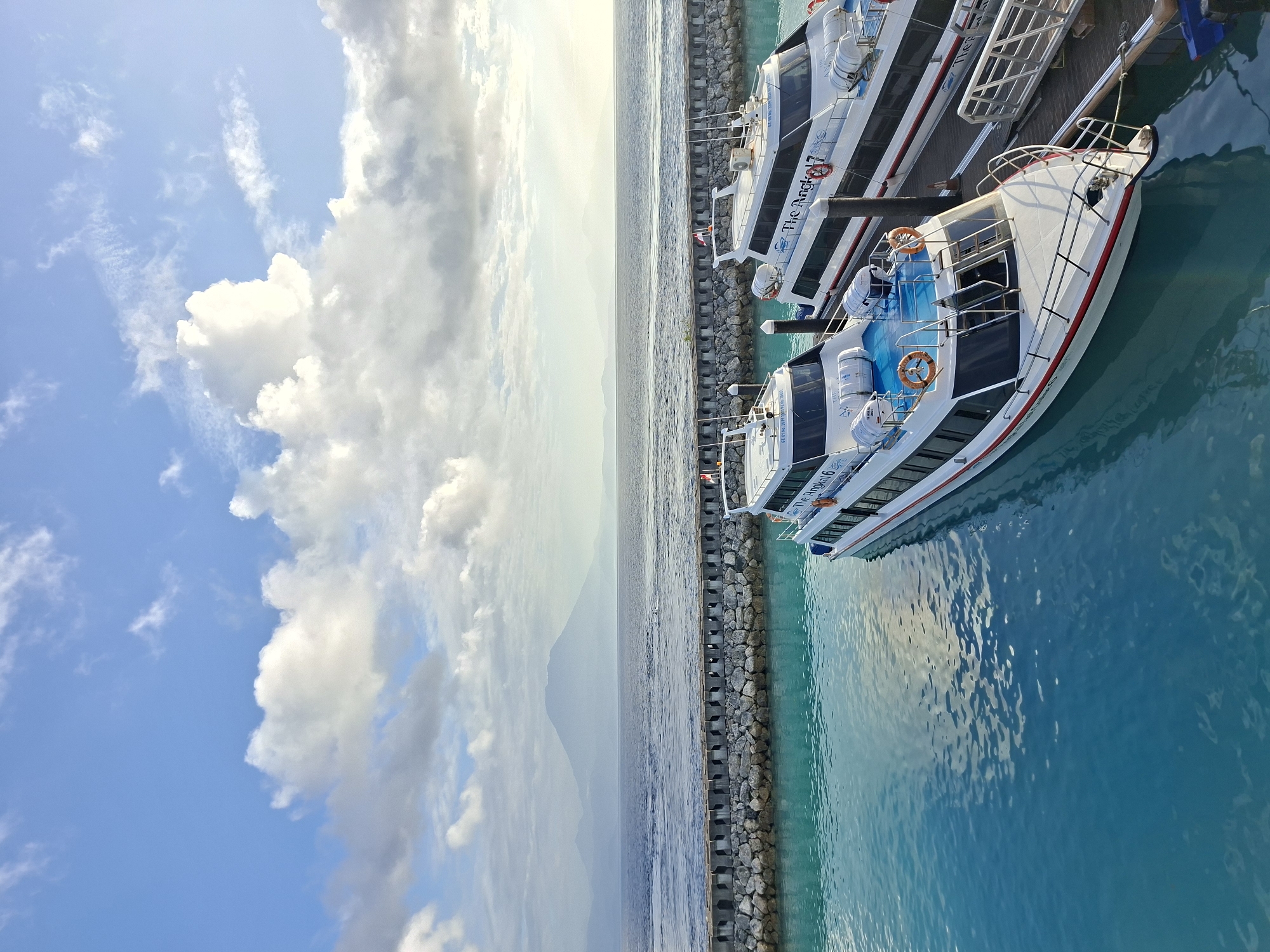 The image shows two white passenger boats docked at a pier on the island of Nusa Penida, Indonesia. The boats, named "The Angkal 6" and "The Angkal 7", are positioned side-by-side. The water in the foreground is a vibrant turquoise color. Behind the pier, the sea stretches to the horizon, where a large, misty mountain range is visible under a partly cloudy sky. The sky is blue with scattered white clouds. The pier is constructed with concrete blocks and rocks. The scene appears to be a harbor or a departure point for sea transportation.