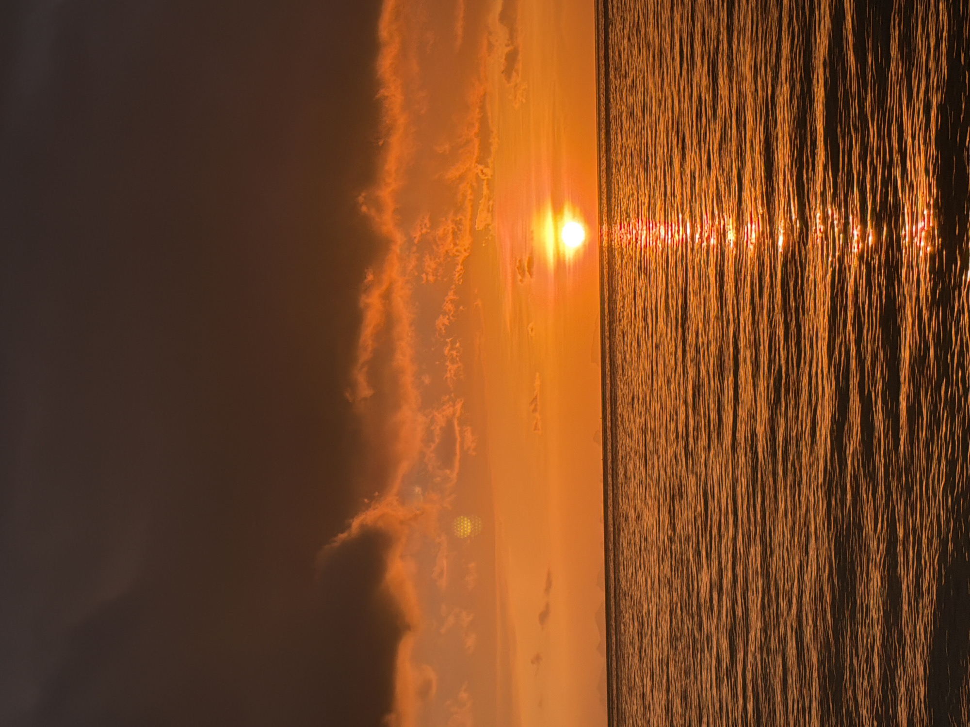 The image depicts a sunset over the ocean in Baan Tai, Thailand. The sky is filled with a gradient of orange and dark red hues, with clouds illuminated by the setting sun. The sun itself is a bright, glowing orb positioned just above the horizon. A reflection of the sun shimmers on the surface of the water, creating a path of light. The sea is calm with gentle ripples. In the distance, the silhouettes of islands or landmasses are faintly visible. The overall scene is one of natural beauty and tranquility.