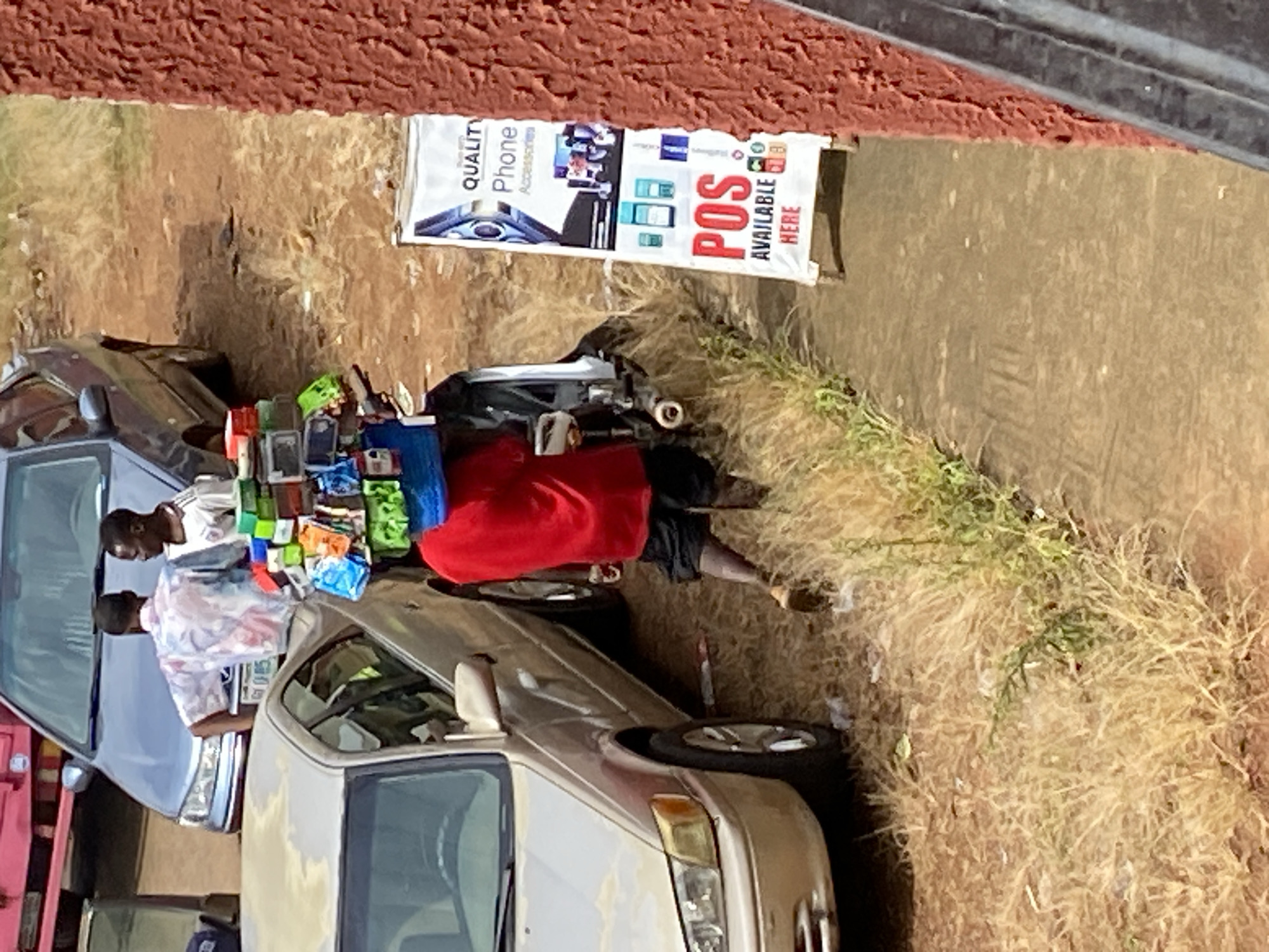This image captures an outdoor street scene in Bukuru, Nigeria. The primary subjects are two men and a variety of objects displayed for sale, along with two cars and a red structure in the foreground.
One man, wearing a patterned white shirt, stands behind a table or display of goods. He appears to be a vendor. Another man, wearing a red garment and black shorts, is also present, possibly a customer or assistant.
The objects for sale are arranged on what appears to be a table or cart. They include numerous phone accessories, such as cases and perhaps chargers, displayed in vibrant colors like green, yellow, blue, and orange. There are also what look like mobile phones themselves. A white banner is visible, advertising "QUALITY Phone Accessories" and "POS AVAILABLE HERE." The banner shows images of phones and payment devices.
Two cars are parked in the scene. One is a silver sedan on the left, and another is a lighter-colored sedan on the right. The ground is covered in dry grass and dirt. The presence of a red structure on the far left suggests a building or stall.
The activity appears to be a street-side vending operation, with the vendor showcasing his phone accessories. The lighting suggests it is daytime, likely outdoors. The setting is consistent with a market or roadside stall in a Nigerian town. The image is slightly tilted, and the perspective is from an elevated viewpoint, looking down on the scene.