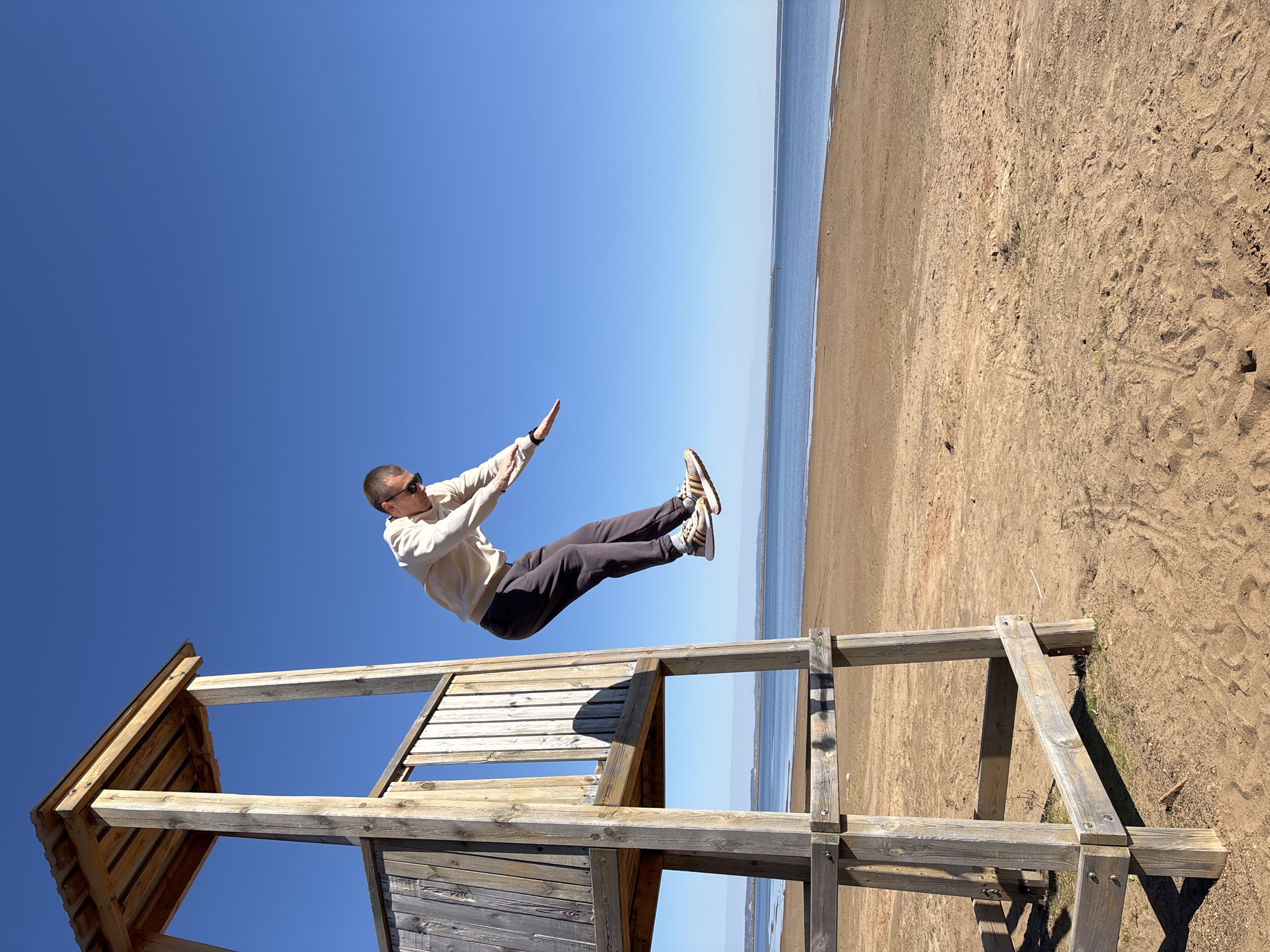A full shot shows a man "jumping" off a lifeguard stand on the coast of Chaikovsky, Russia. He is wearing sunglasses, a light-colored sweatshirt, and dark pants, and he has both arms raised. The lifeguard stand is made of wood and has a partially enclosed top section. The coast consists of a sandy beach and blue water with a light horizon, and the sky is clear blue. The image is rotated so that the beach is on the right.