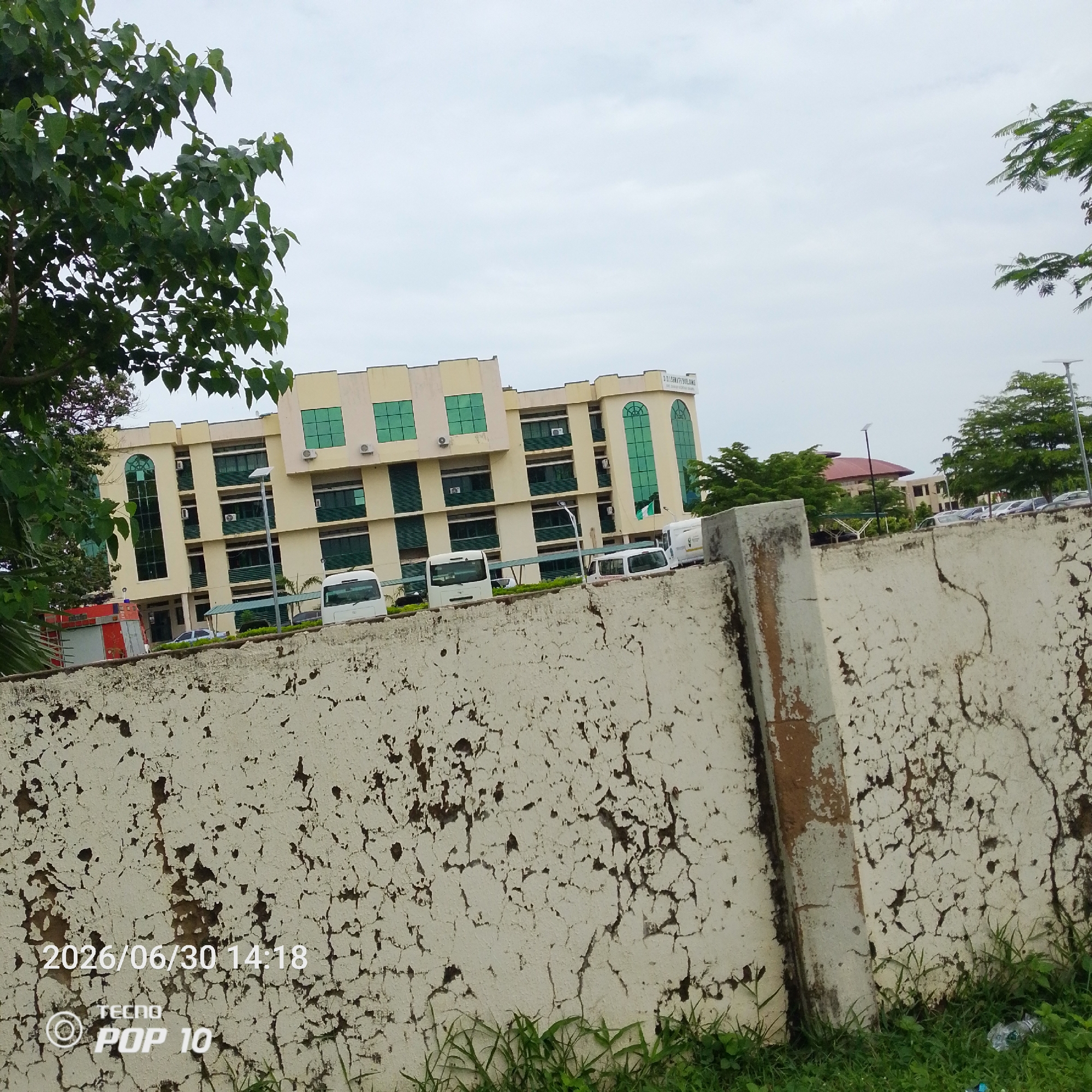 This is a medium shot taken outdoors in Jalingo, Nigeria. The scene features a modern, multi-story building with cream-colored walls and green-tinted windows. The building appears to be an office or institutional structure, possibly with commercial spaces on the ground floor, as suggested by a red vehicle that resembles a fire truck parked to the left. Several white vans or minibusses are parked in front of the building, and cars are visible further in the background, indicating a likely parking lot or street.
A rough, textured concrete wall with peeling paint dominates the foreground, running diagonally across the image. The wall is partially covered in green grass and weeds at its base, suggesting an overgrown or naturalized area. Tall trees with dense green foliage frame the top left and right of the image, and the sky above is overcast with a pale, cloudy appearance.
A date and time stamp are visible in the bottom left corner: "2026/06/30 14:18". Below this, there is text that reads "TECNO POP 10", likely indicating the device used to capture the photo. The overall mood of the image is neutral, conveying a sense of an ordinary day in an urban or semi-urban environment. The weather appears to be overcast, which might suggest a humid or potentially rainy climate typical of Jalingo. There are no people clearly visible in the frame, focusing the attention on the architectural and environmental elements of the scene.