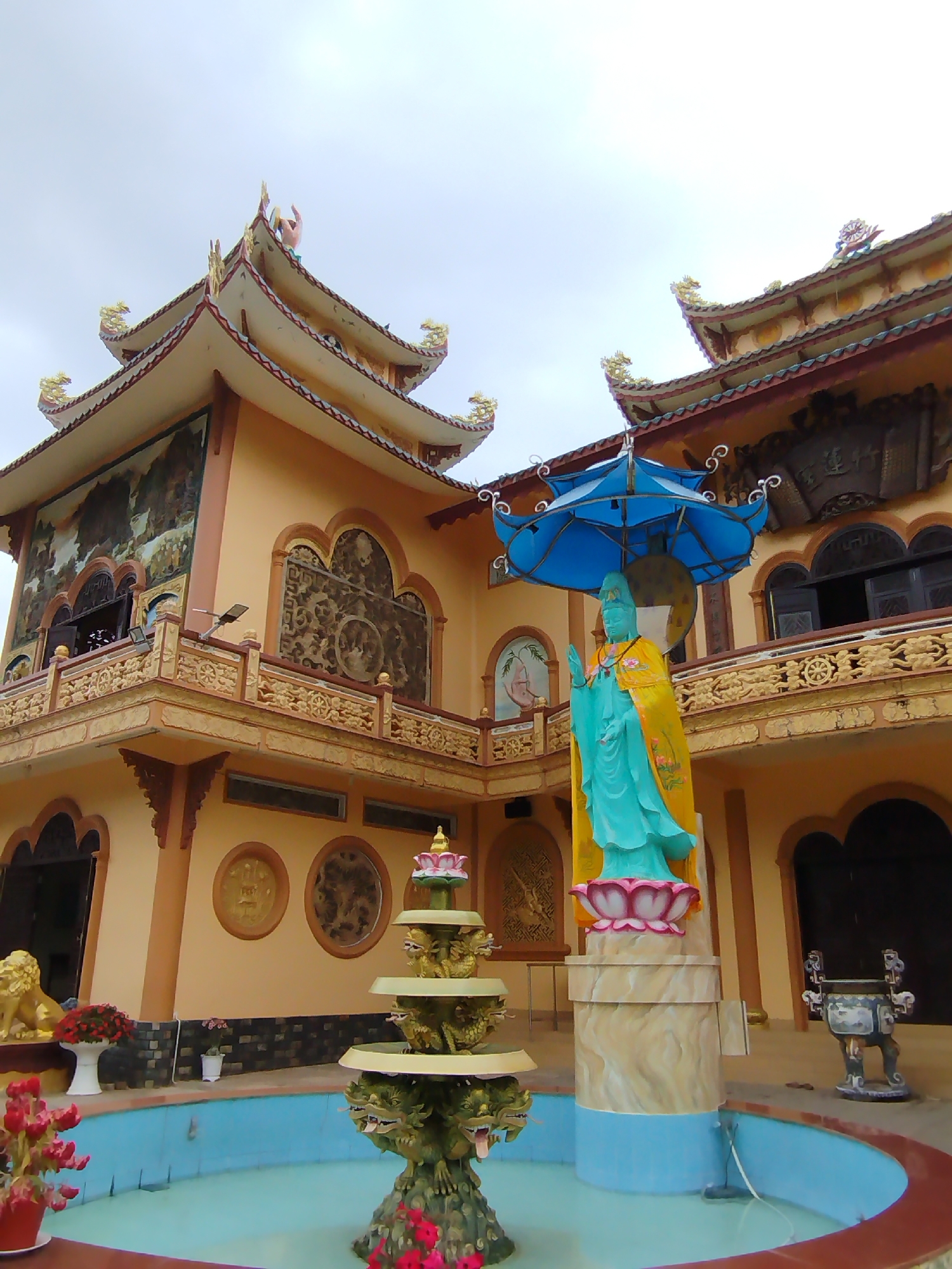 The image depicts a temple in Phú An, Vietnam. The temple features a tan-colored multi-tiered structure with ornate carvings and murals on the exterior walls. A statue of a figure with a teal robe is positioned in front of the building, sheltered by a blue umbrella. In the foreground, a tiered fountain sits in a blue pool. Other statues, flowers, and an incense burner are also visible. The sky is overcast.